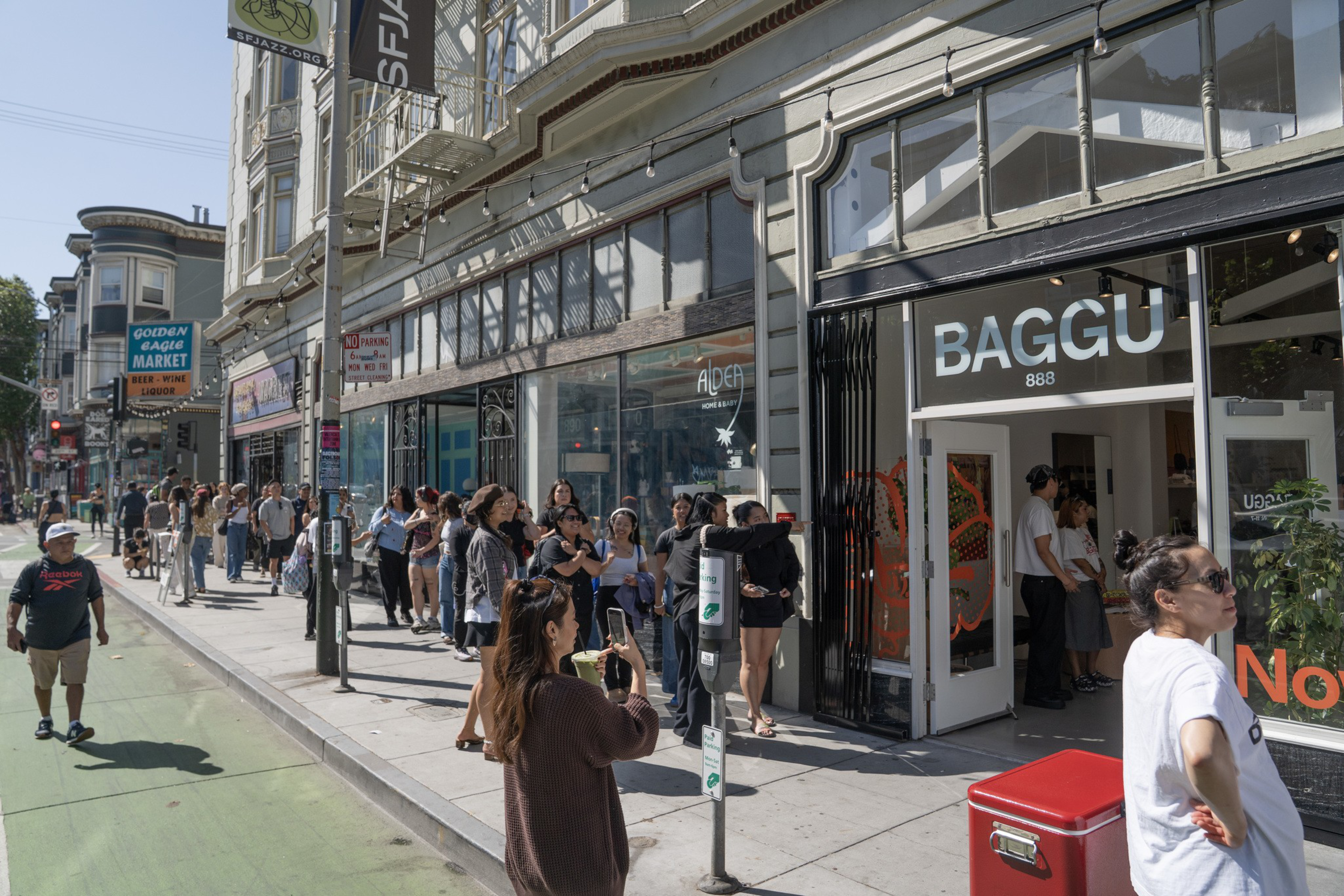 A long line of people waits outside the BAGGU store on a sunny city street, while others walk or stand nearby.