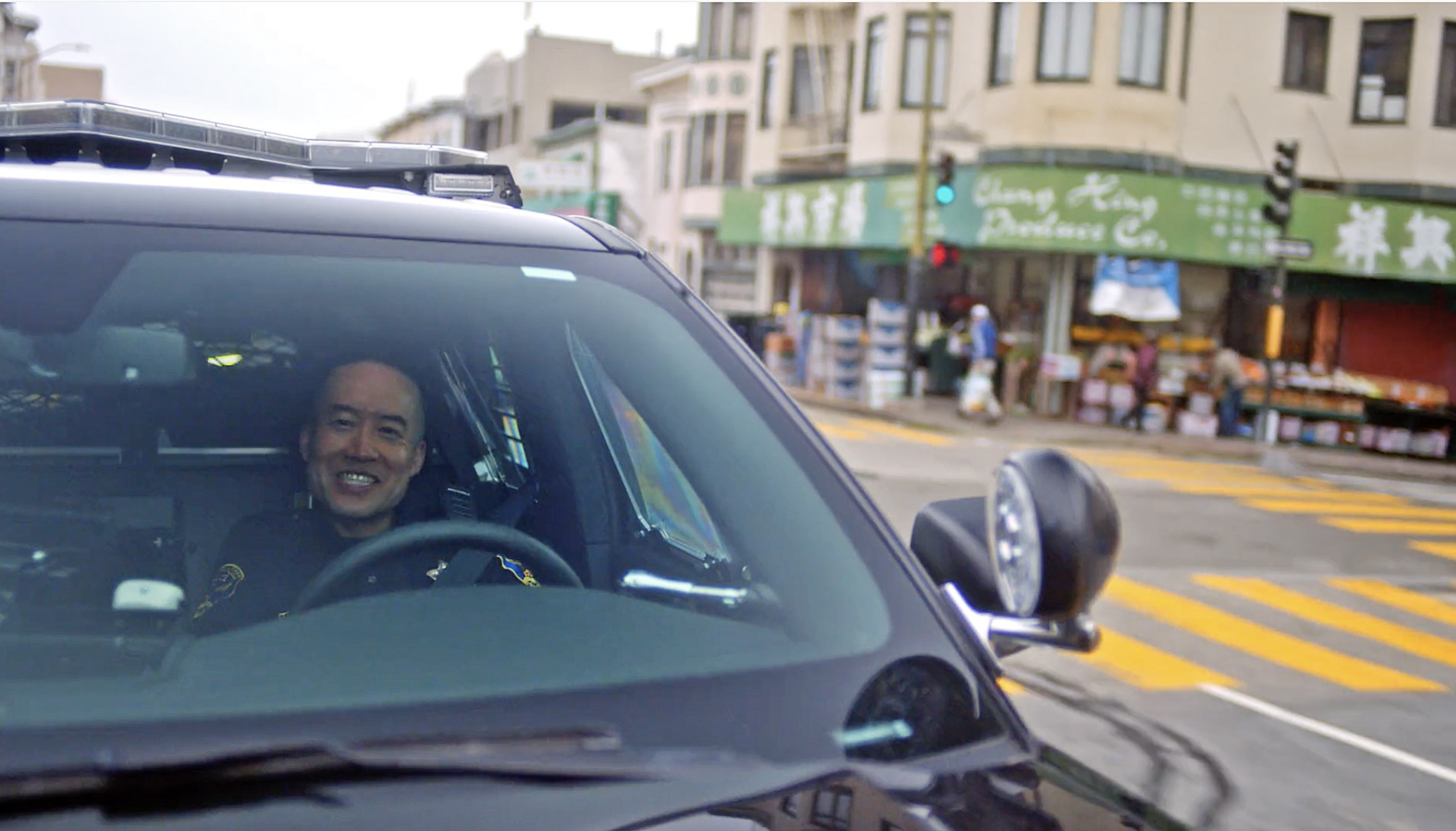 A smiling police officer sits inside a black police SUV stopped at a street intersection with yellow crosswalk lines and shops in the background.