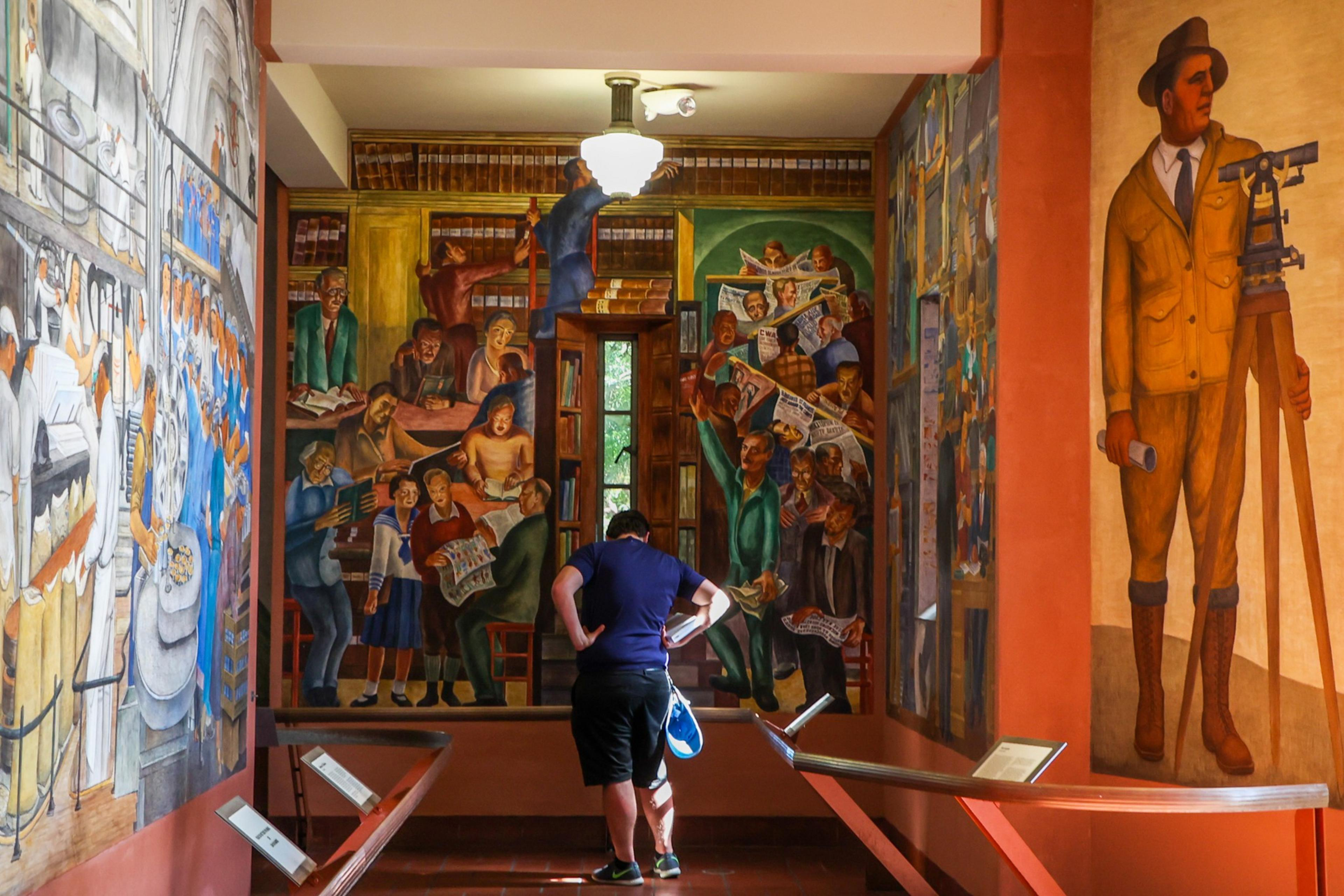 A person stands in front of vibrant murals depicting groups of people reading newspapers and a man with surveying equipment.