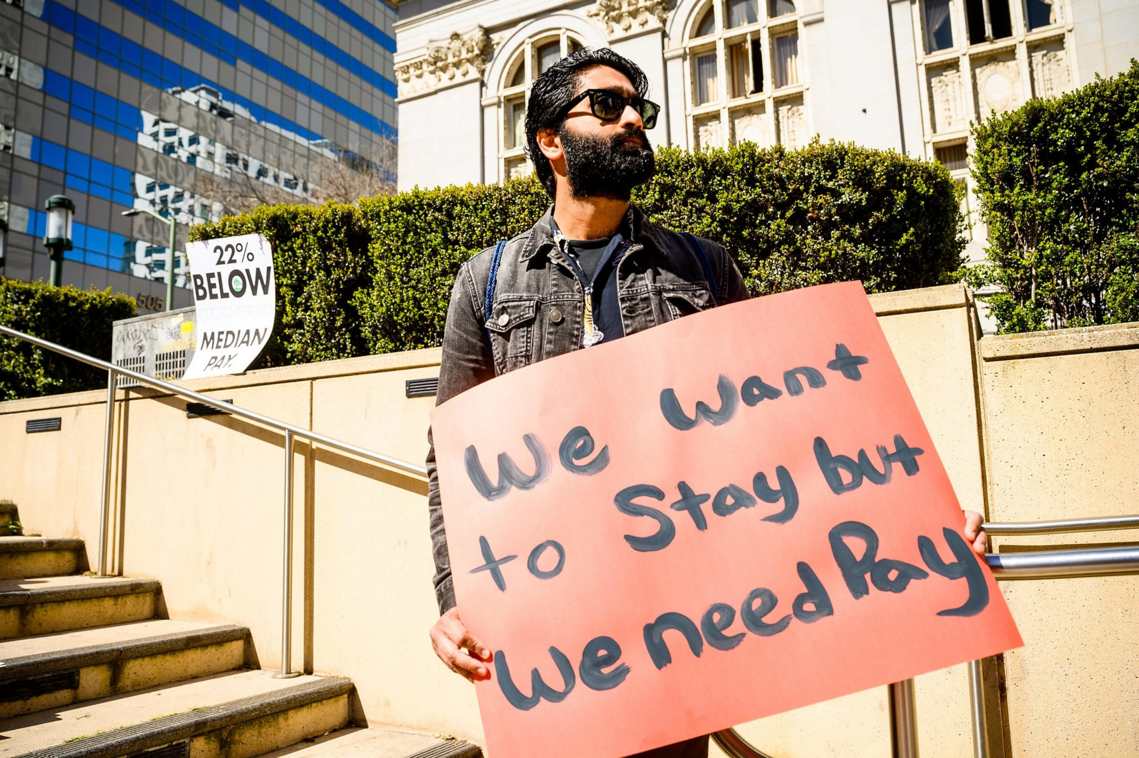 A bearded man wearing sunglasses holds a sign that says, "We want to stay but we need pay," standing outdoors near steps and buildings.