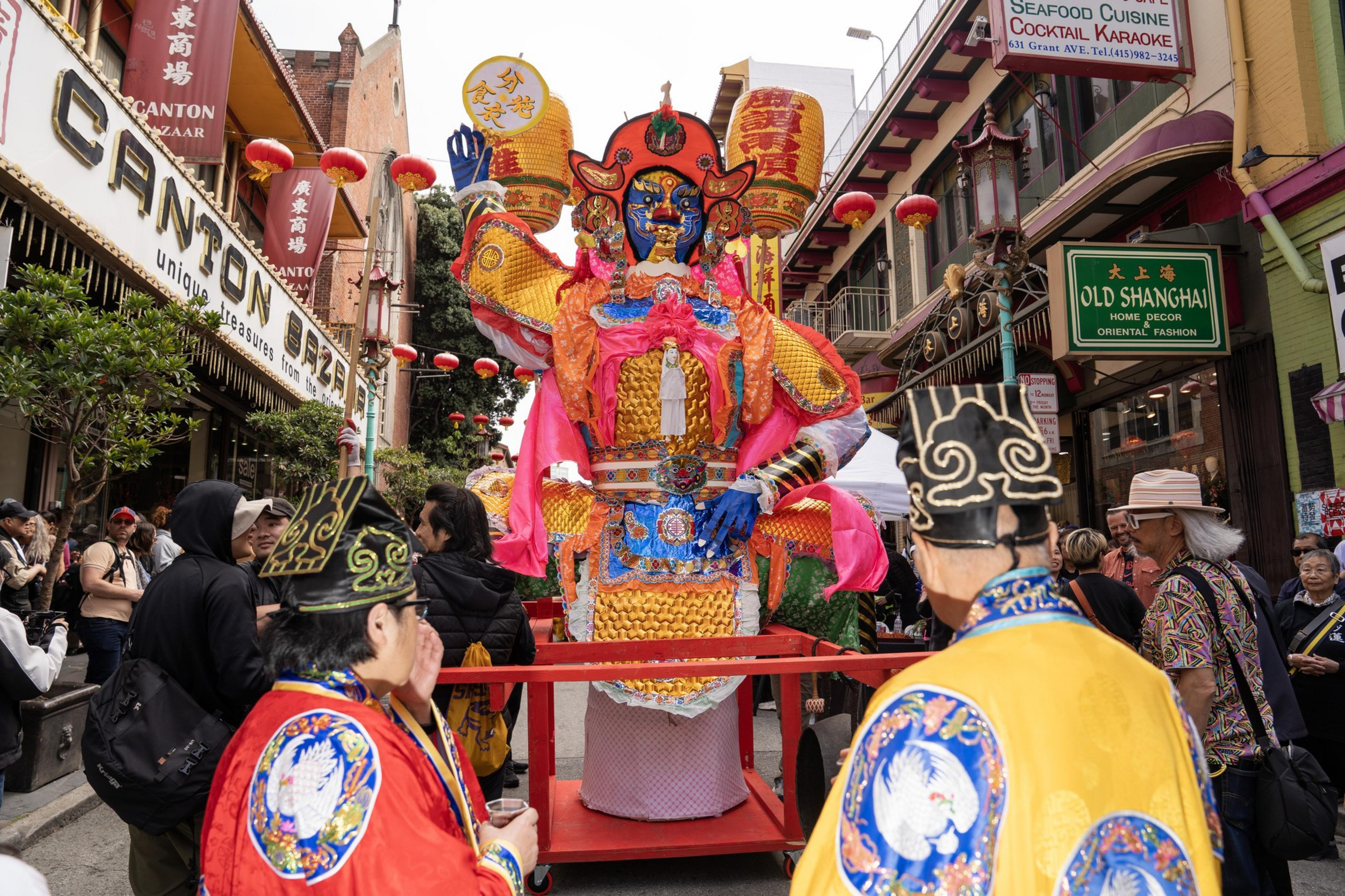 A vibrant Chinese parade features a large, ornate figure in bright orange and pink, surrounded by people in traditional costumes and lanterns hanging above.