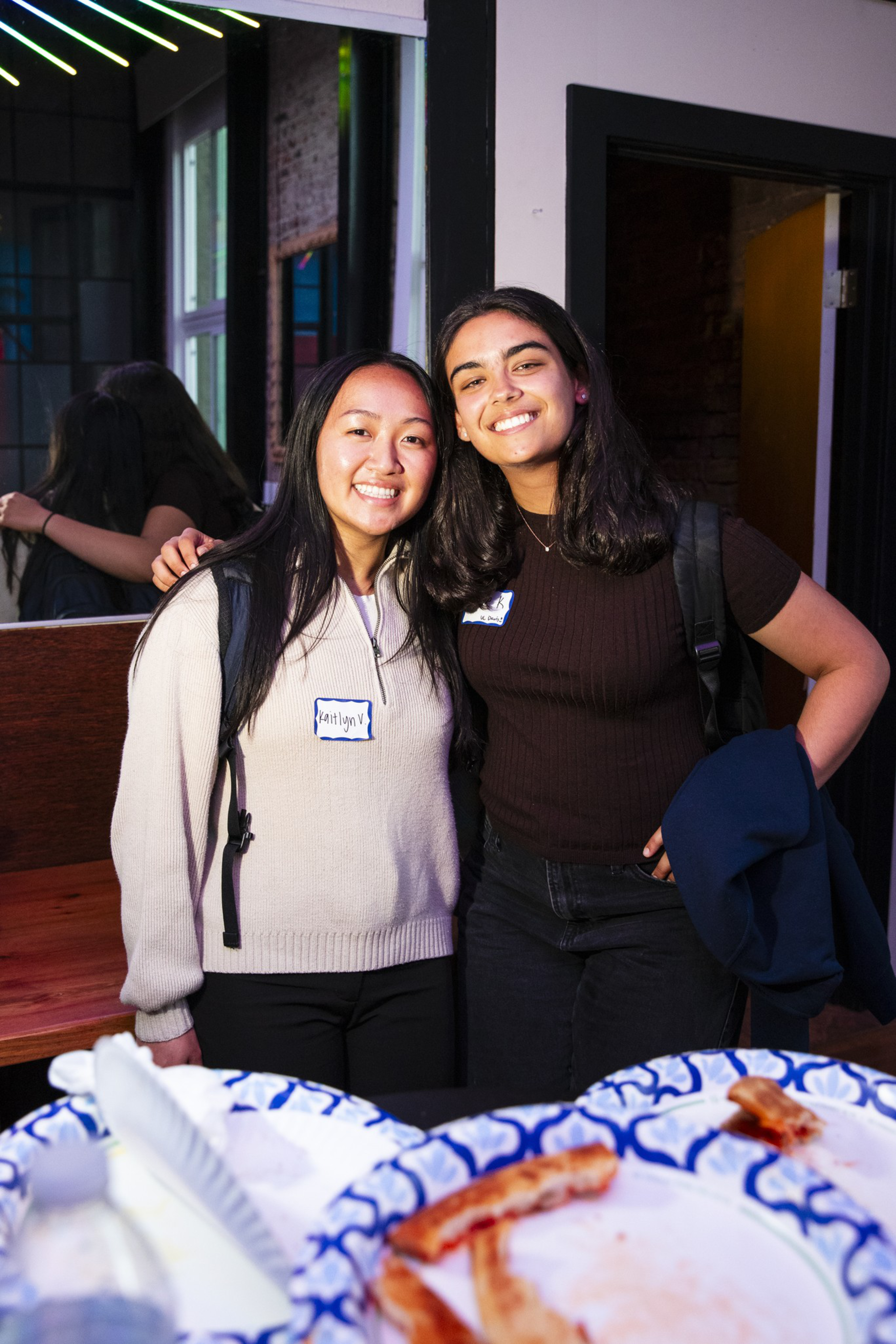 Two smiling women stand close together, wearing name tags and backpacks, behind plates with leftover ribs on a table.