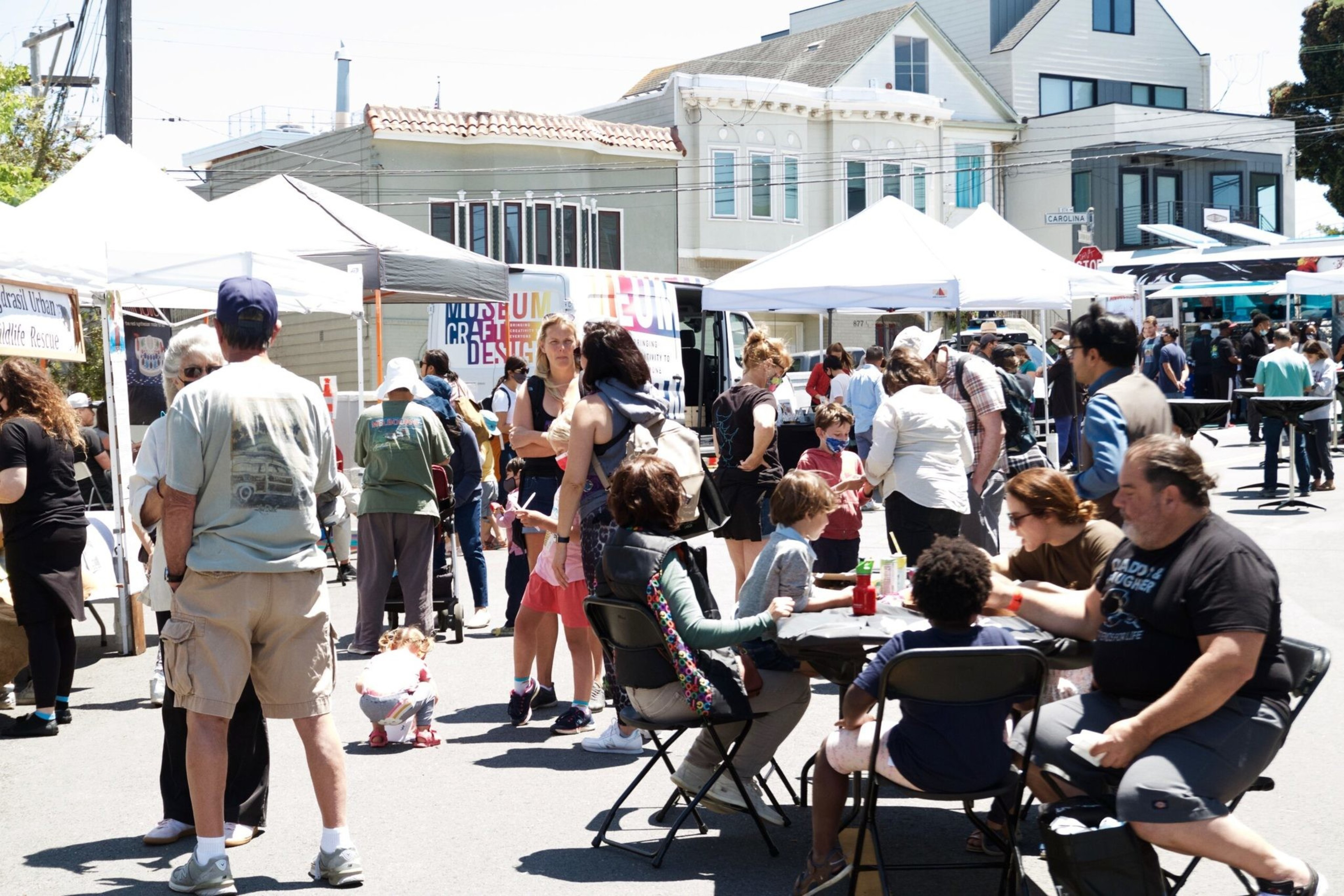 People are walking, talking, and sitting at tables under white tents on a sunny day at an outdoor street market or fair.