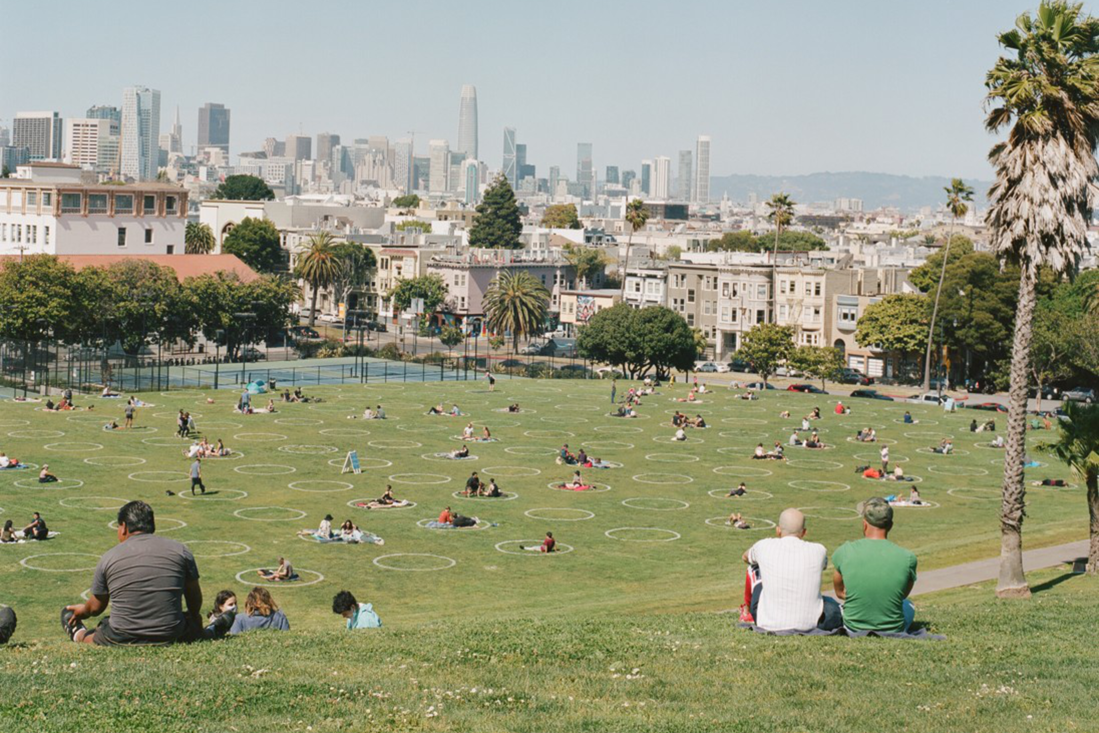 People are enjoying a sunny day in a spacious park, sitting in distanced circles on the grass. The city skyline is visible in the background.