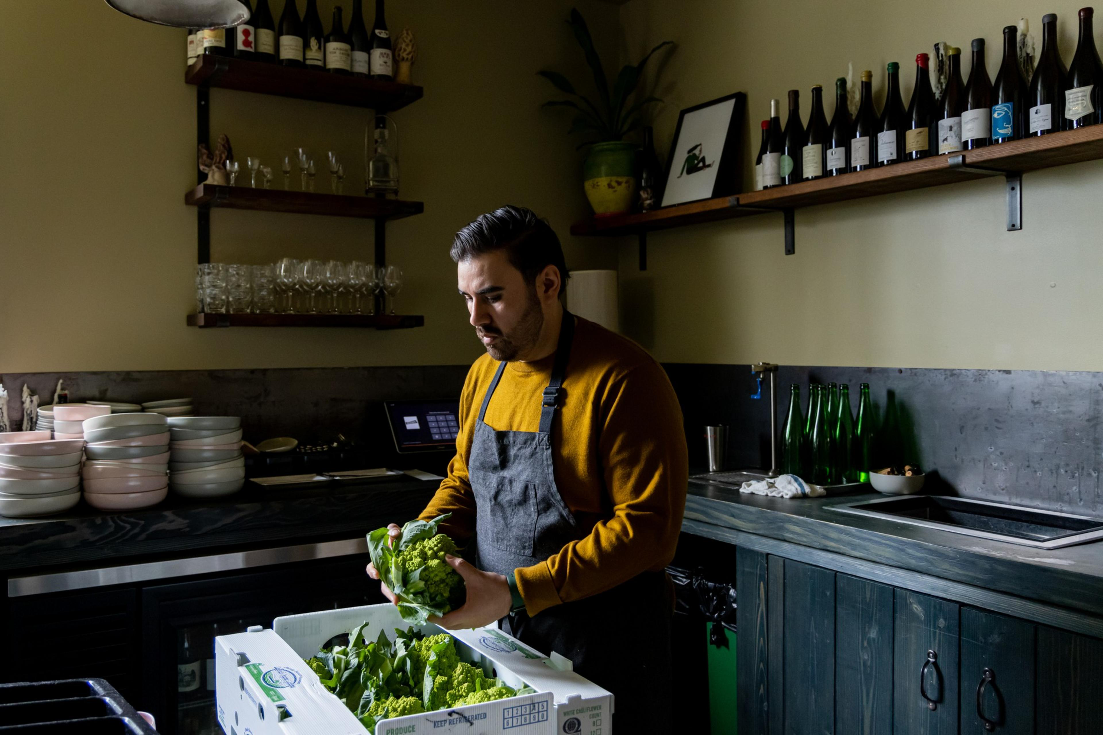 A man in a mustard sweater and apron holds a leafy vegetable in a cozy kitchen with shelves of wine bottles and glasses around him.