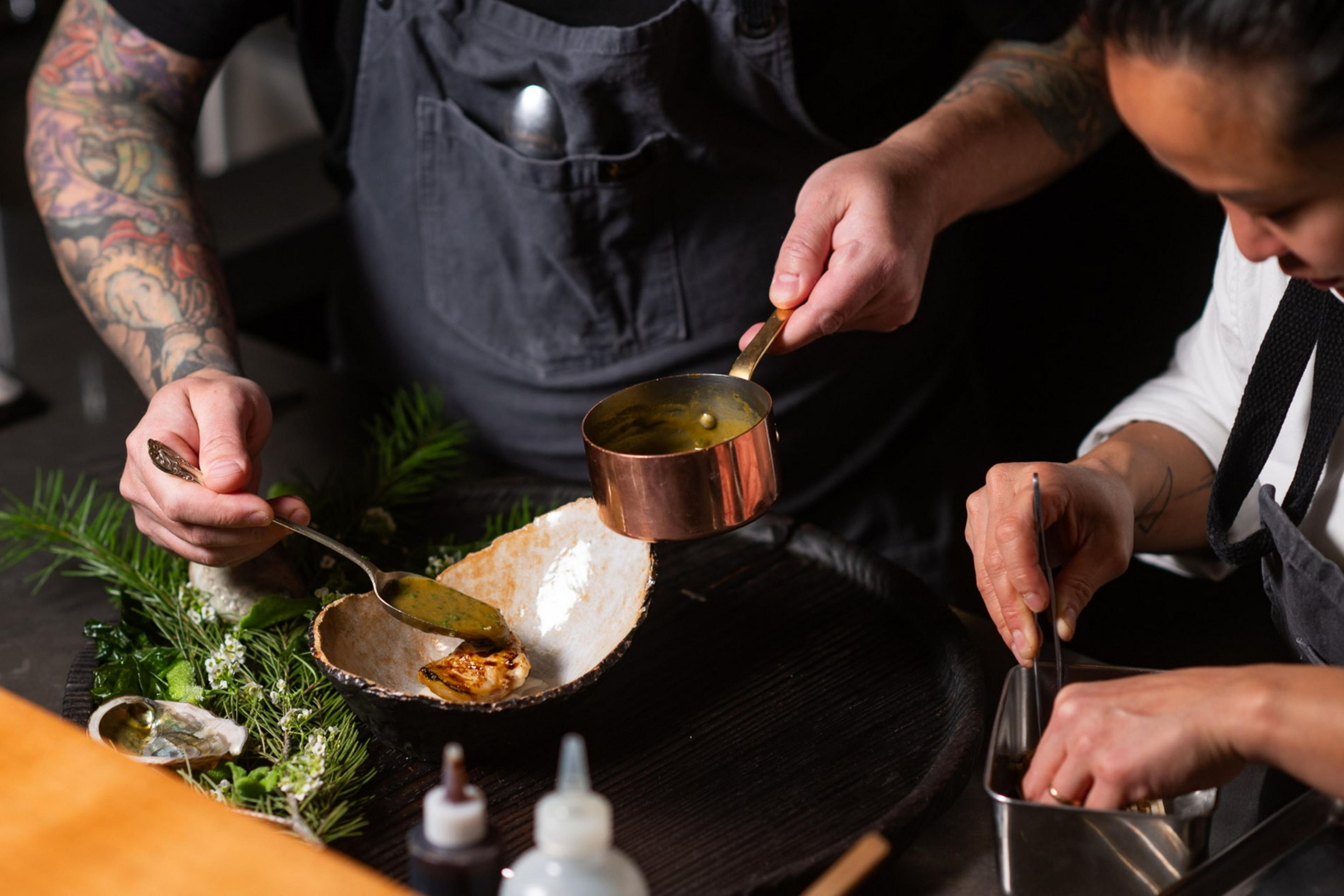 Two chefs prepare a dish, carefully spooning sauce from a copper pot onto a plate with greens and seafood. They wear aprons and have tattoos.
