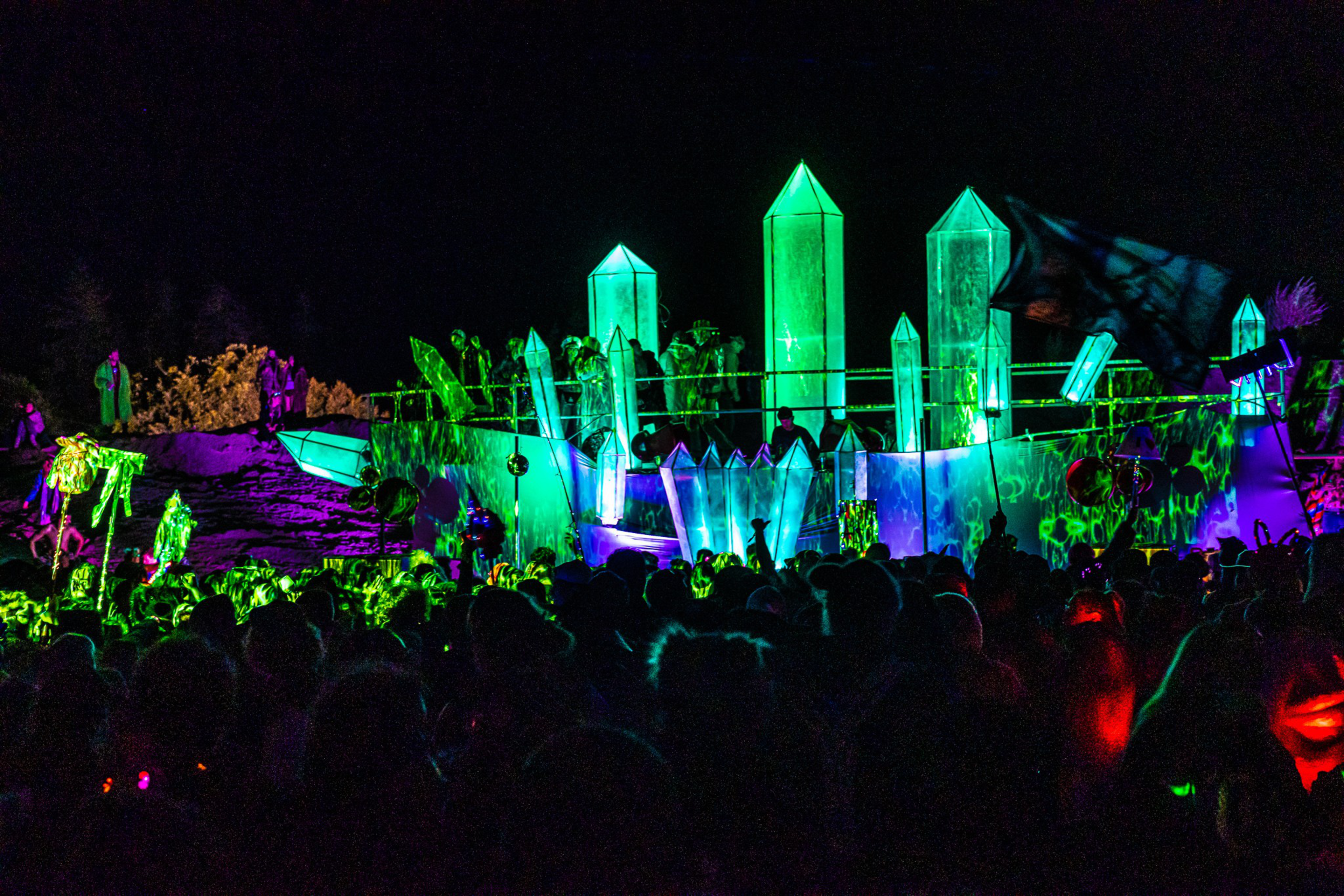 A crowd gathers at night around glowing green and blue crystal-shaped light sculptures with people standing atop the installation.