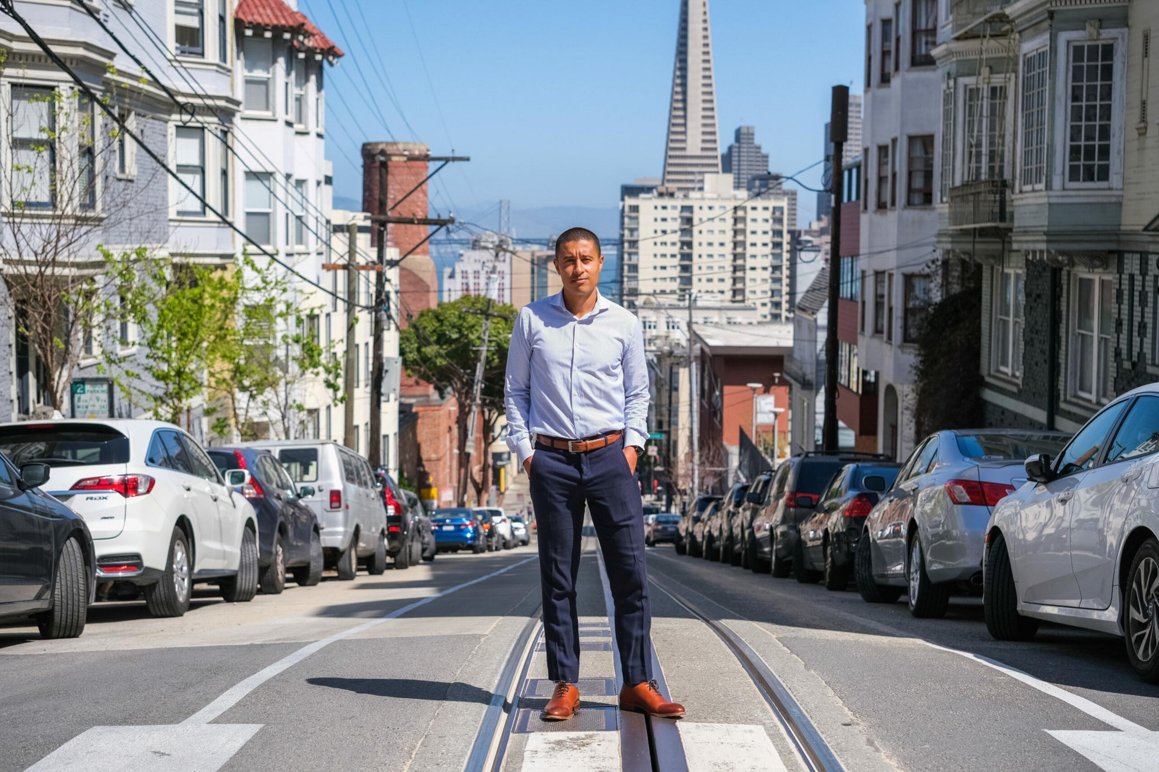 A man in a light blue shirt and dark pants stands confidently on a cable car track in a sunny urban street with parked cars and tall buildings.