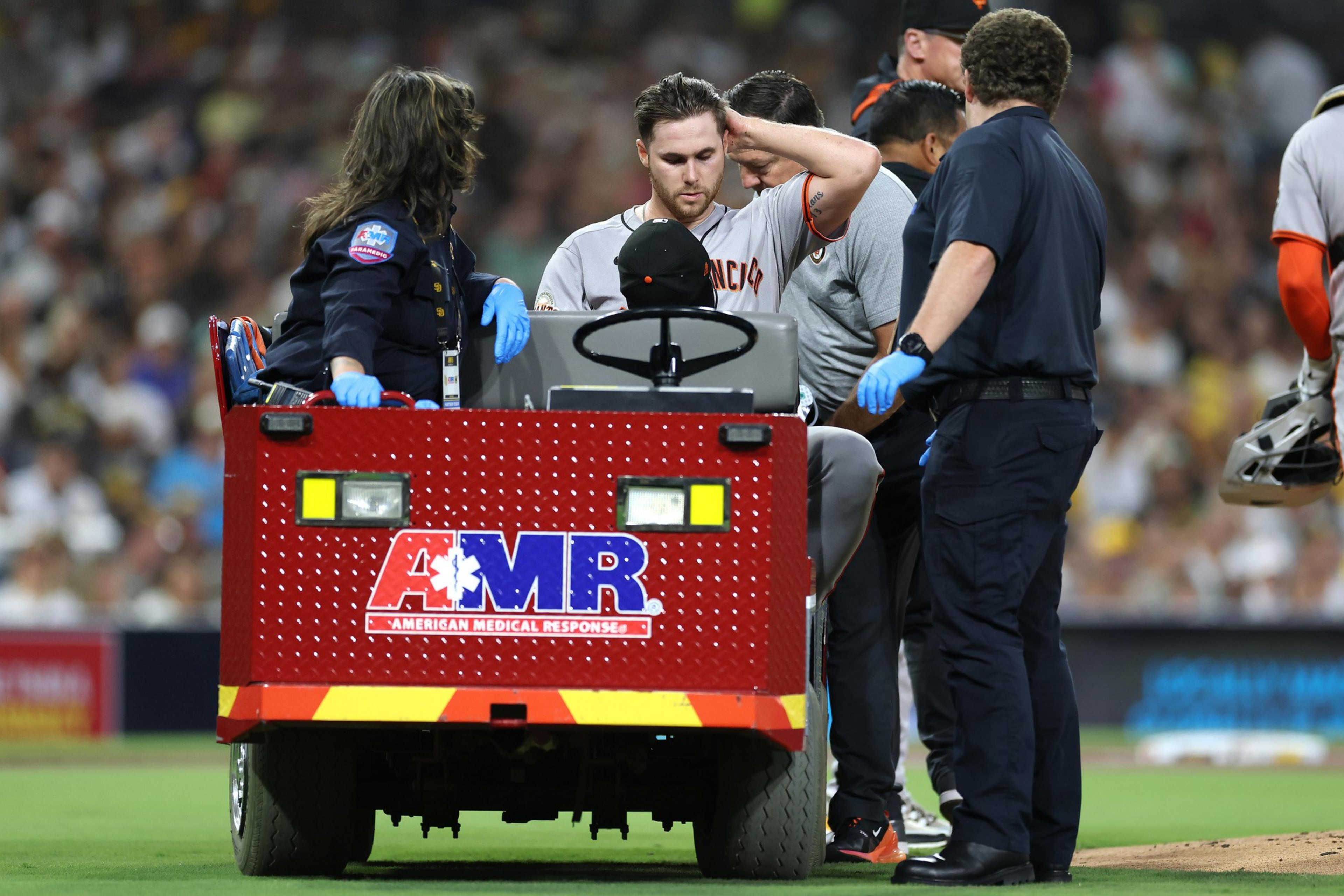 A baseball player in a San Francisco uniform sits on a medical response cart surrounded by three medics wearing gloves during a game.