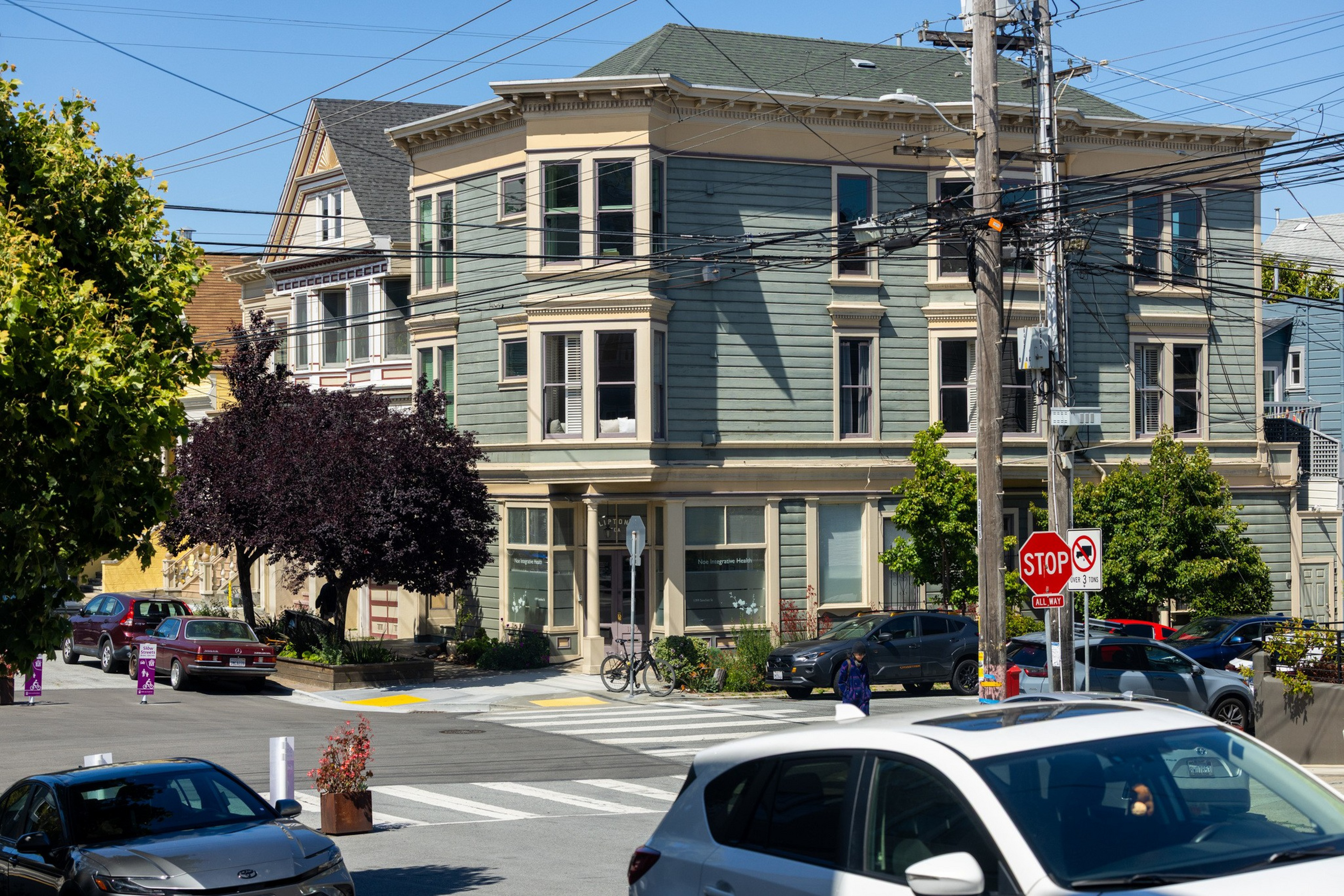 A green three-story building sits on a sunny street corner with cars parked along the road and people walking nearby.