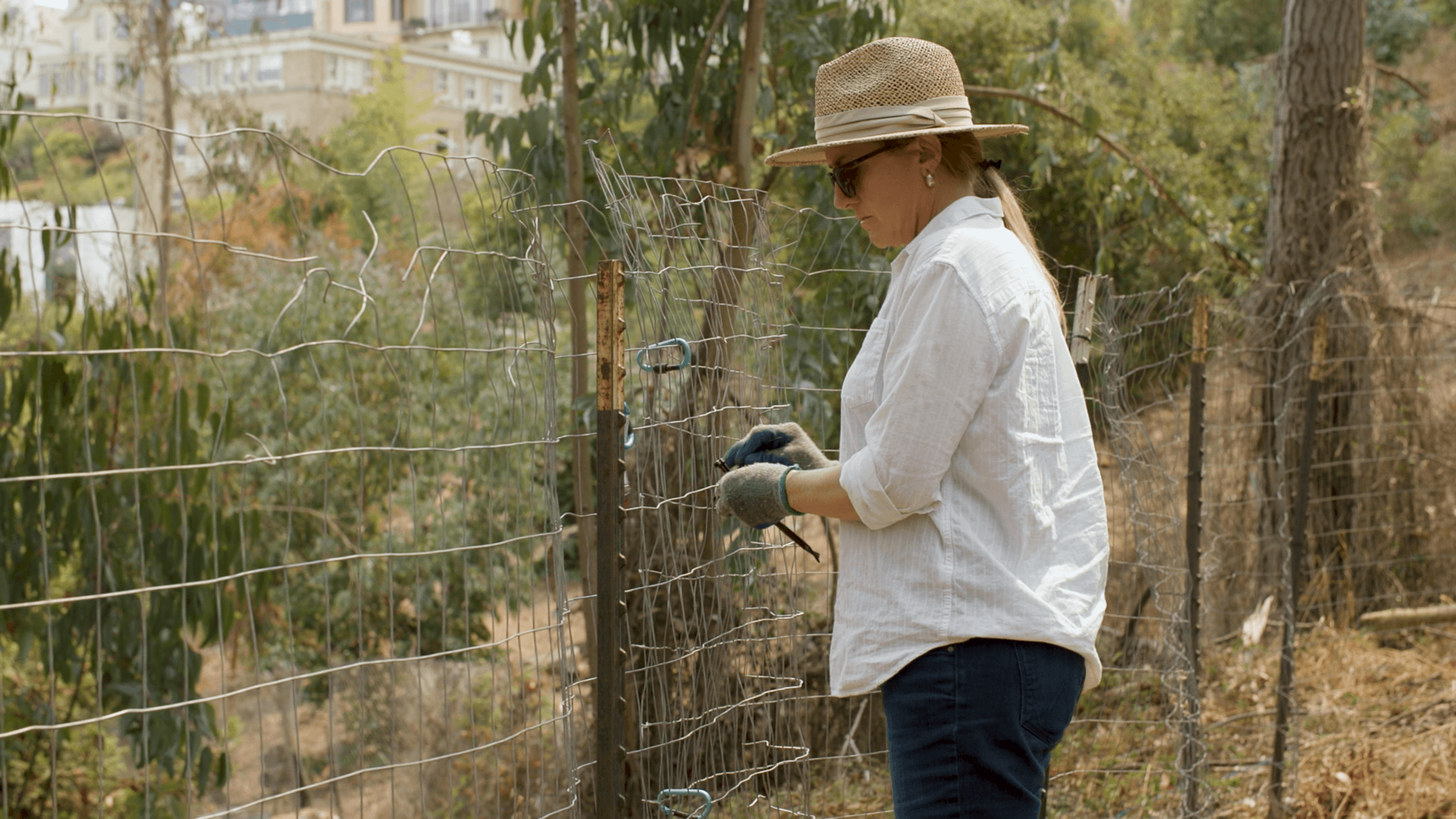 A woman wearing a straw hat, sunglasses, white shirt, and gloves repairs a wire fence in a dry, wooded area with buildings in the background.