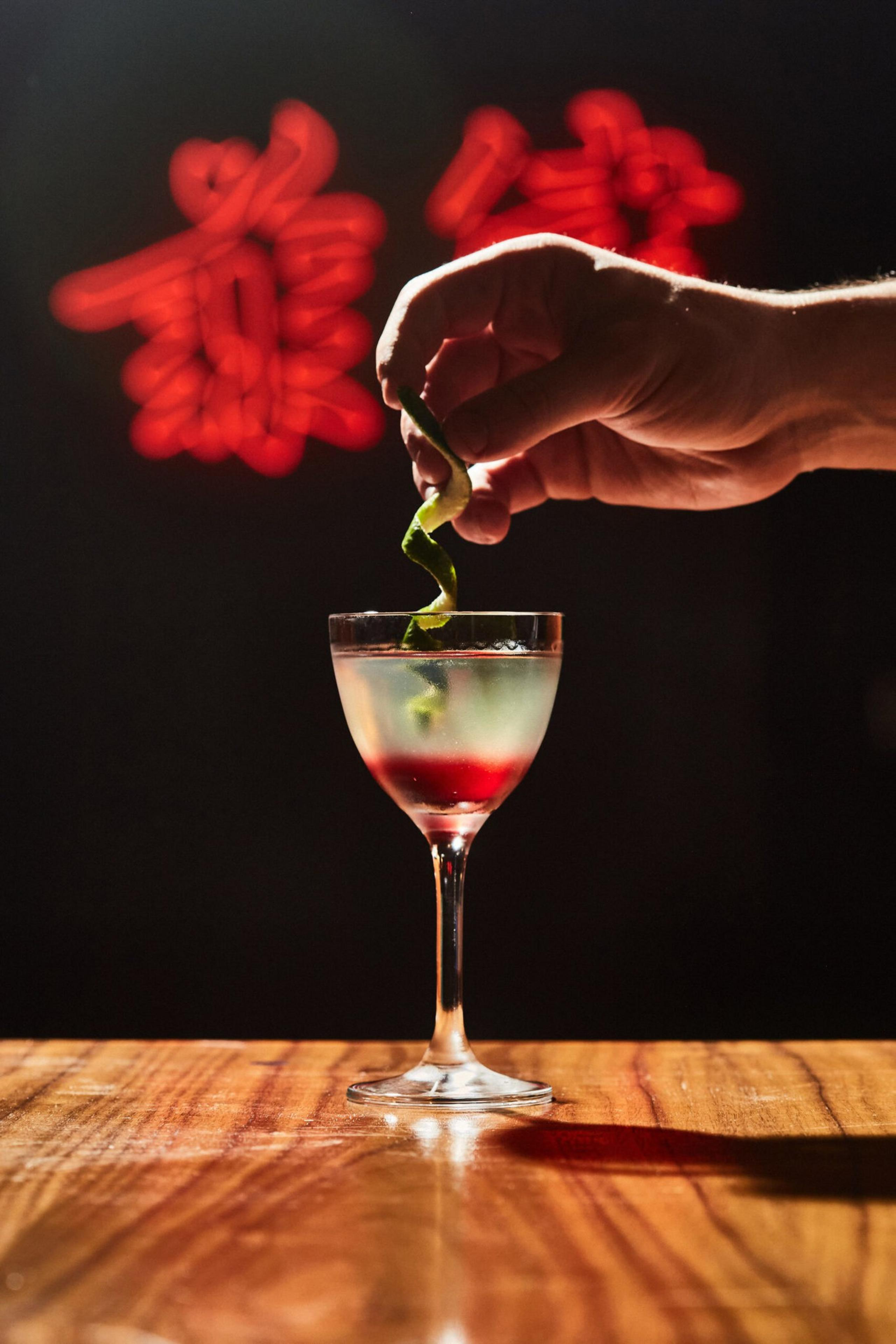 A hand twists a green peel garnish over a cocktail glass with a layered red and clear drink, set on a wooden surface with red neon characters in the background.