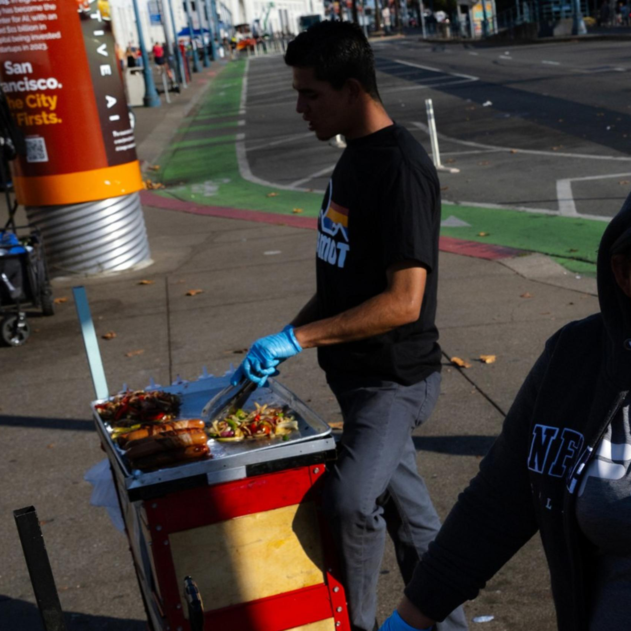 A woman in a hoodie stands next to a food cart with grilled sausages and onions. Another person is preparing food in the background on a city street.