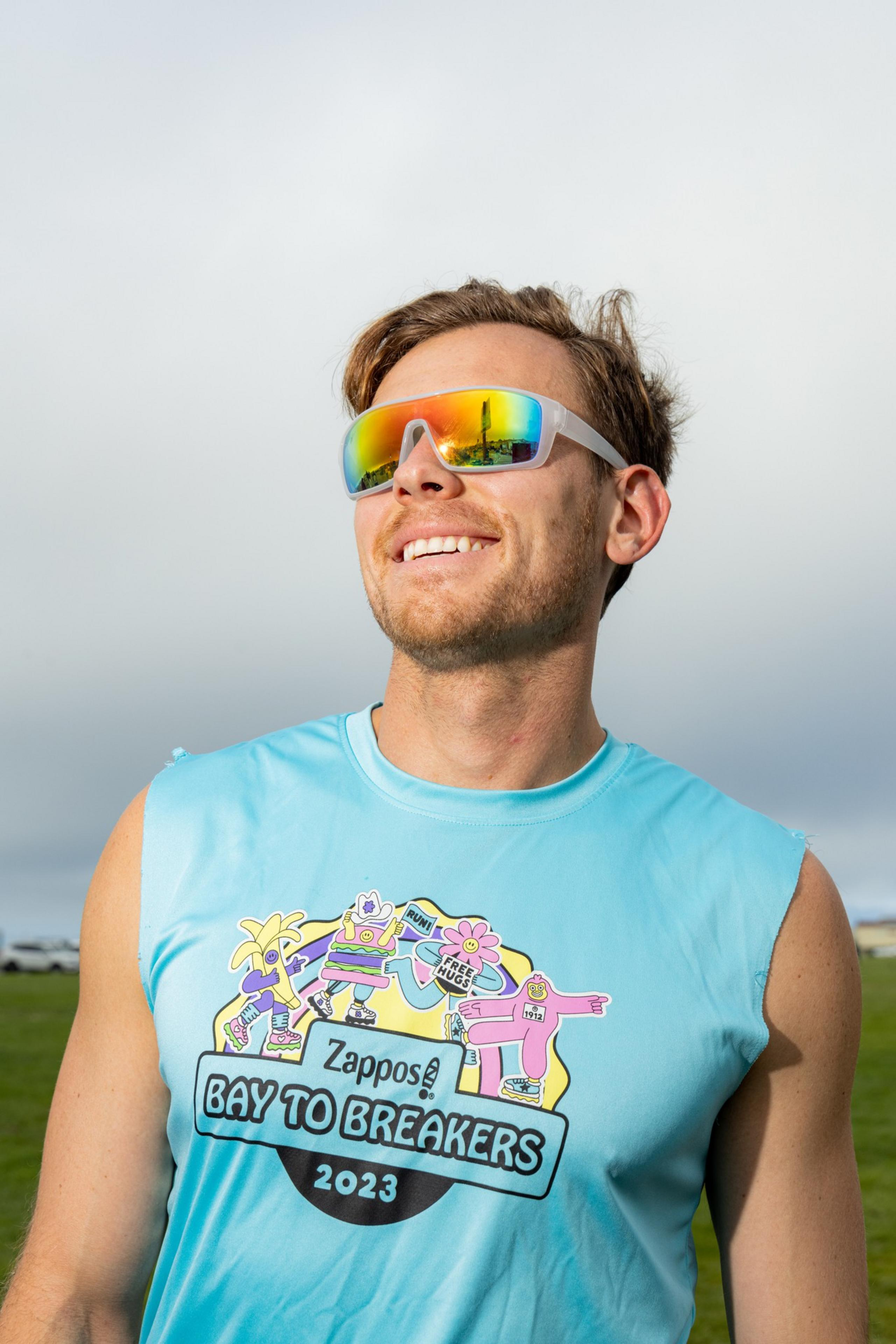A person is smiling, wearing reflective rainbow sunglasses and a sleeveless blue shirt that says &quot;Zappos Bay to Breakers 2023&quot; with colorful cartoon characters on it.