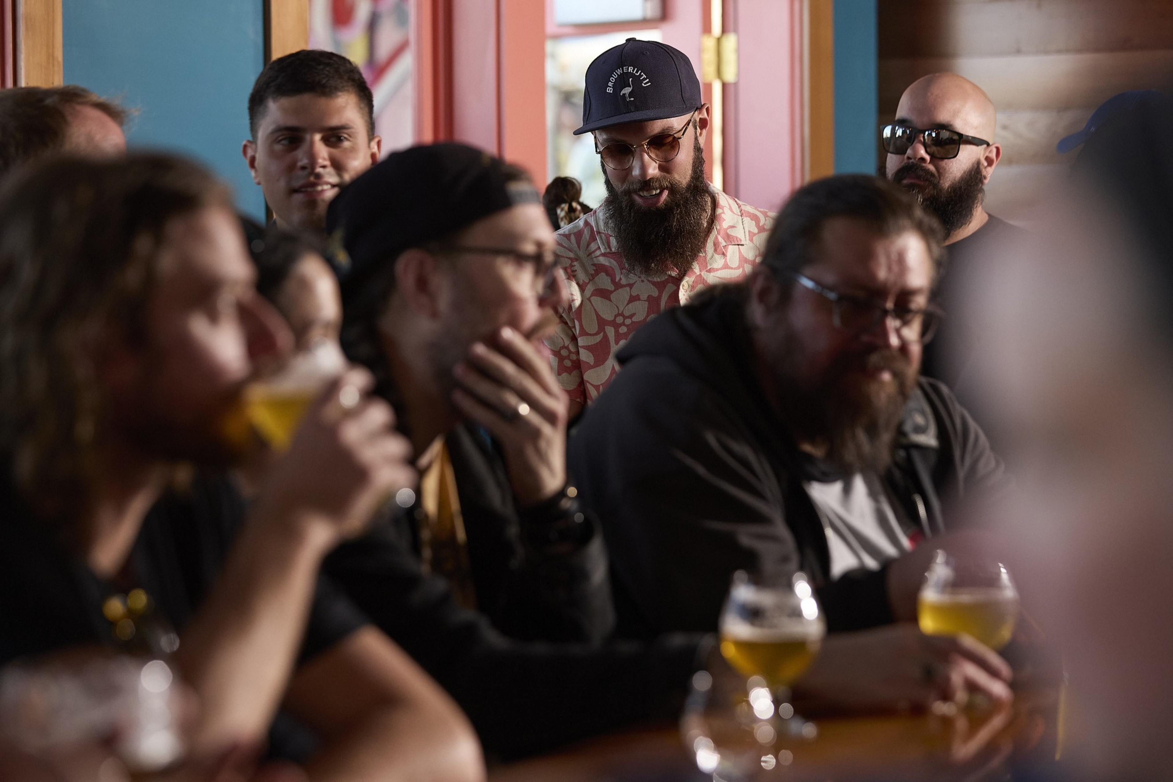 A group of men are gathered indoors, some holding glasses of beer. They are casually dressed, and the scene is in a lively, social setting with a warm atmosphere.
