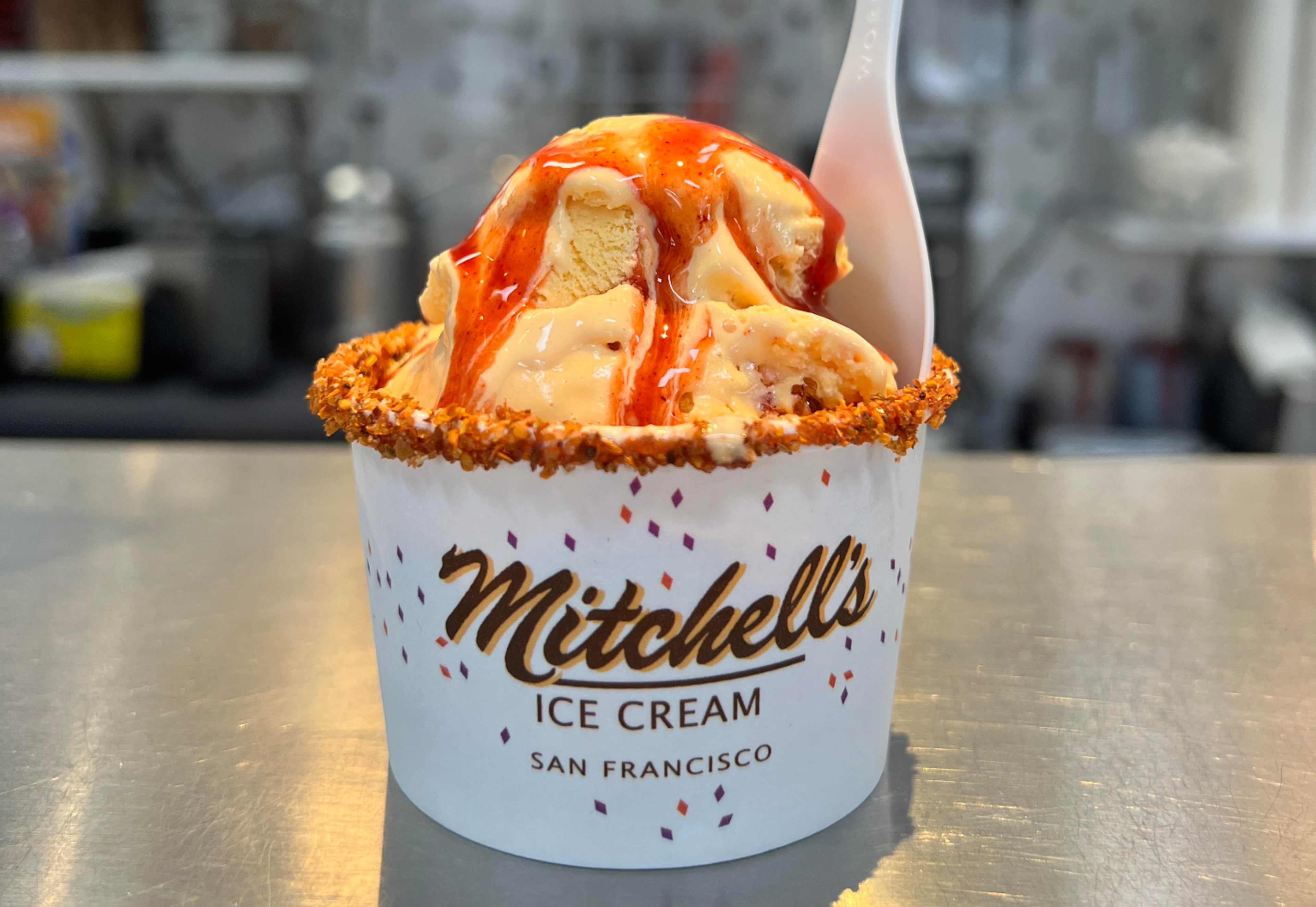 A cup of Mitchell's ice cream, topped with red syrup and surrounded by a crunchy rim, sits on a countertop with a white spoon tucked inside.