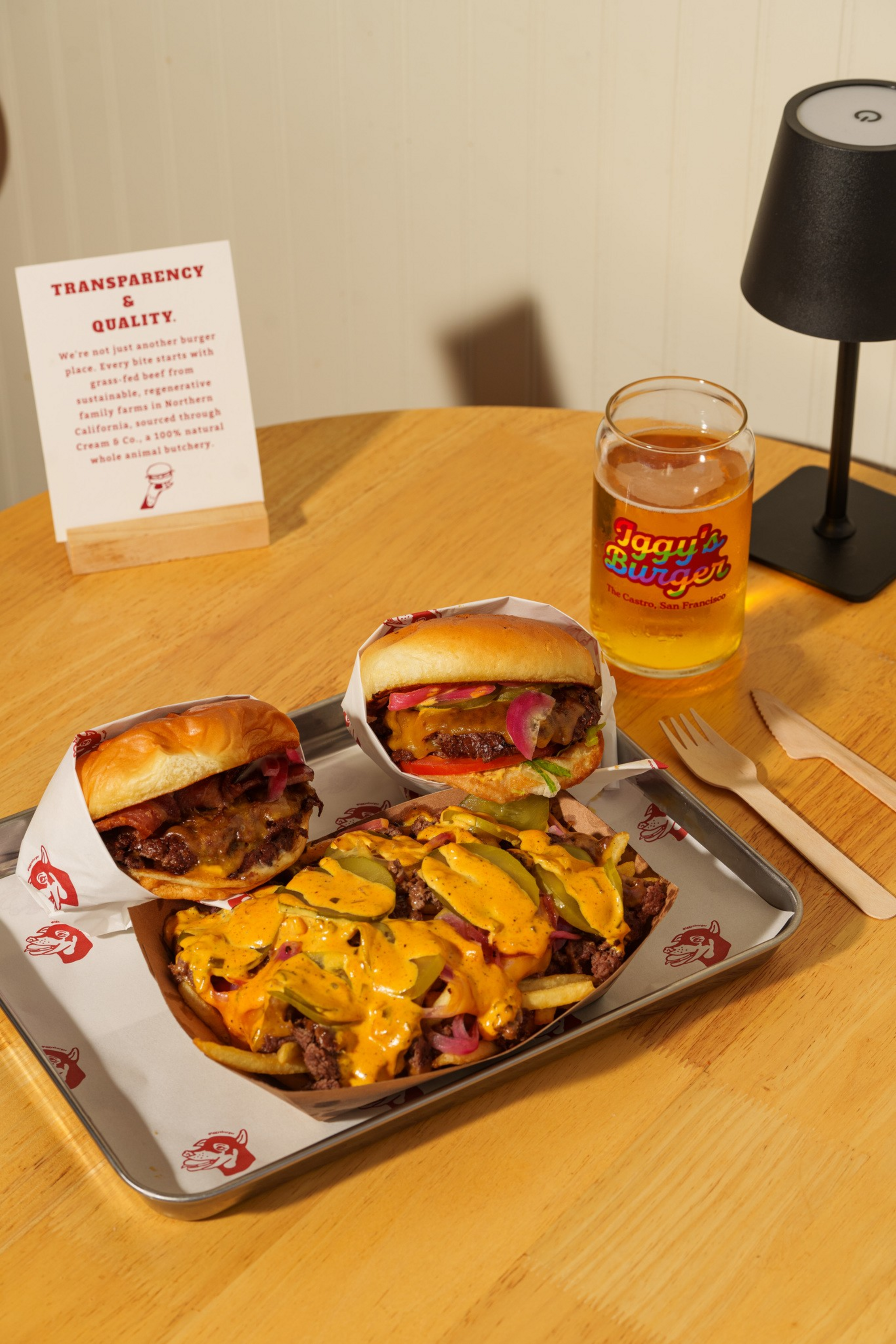 Two cheeseburgers and a tray of loaded fries with cheese and pickles sit on a wooden table next to a branded glass of beer and a small lamp.