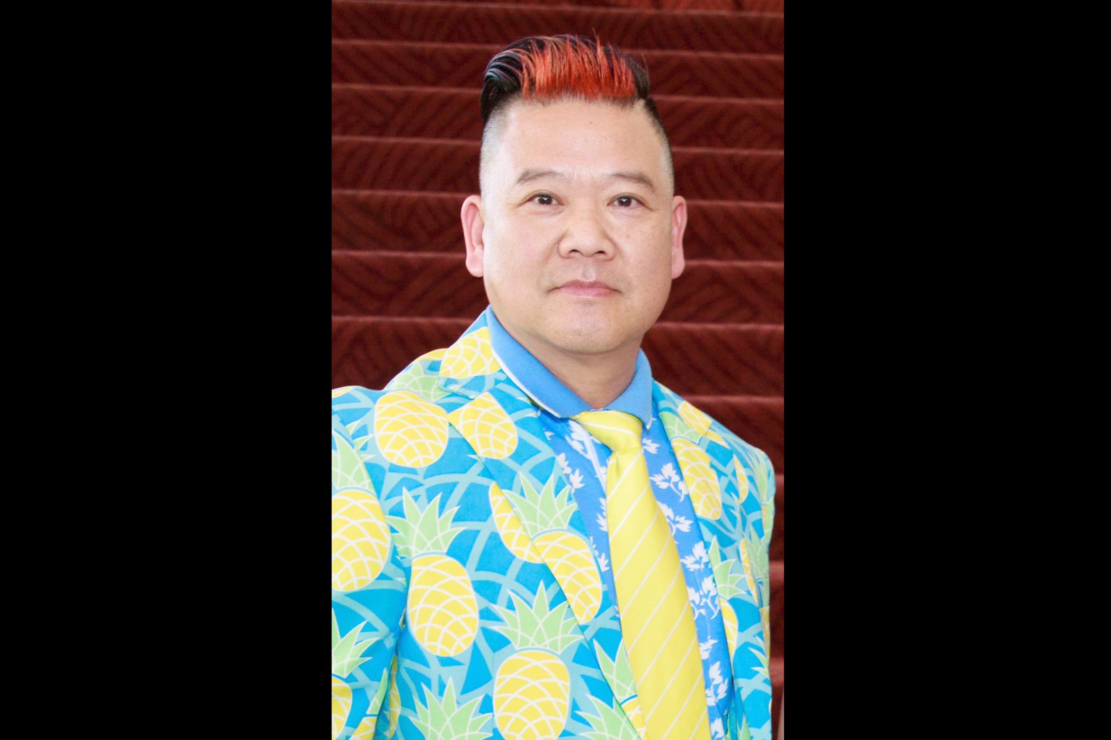 A man wears a blue suit covered in large yellow pineapples, a blue shirt, and a bright yellow tie, with short hair dyed orange at the front.