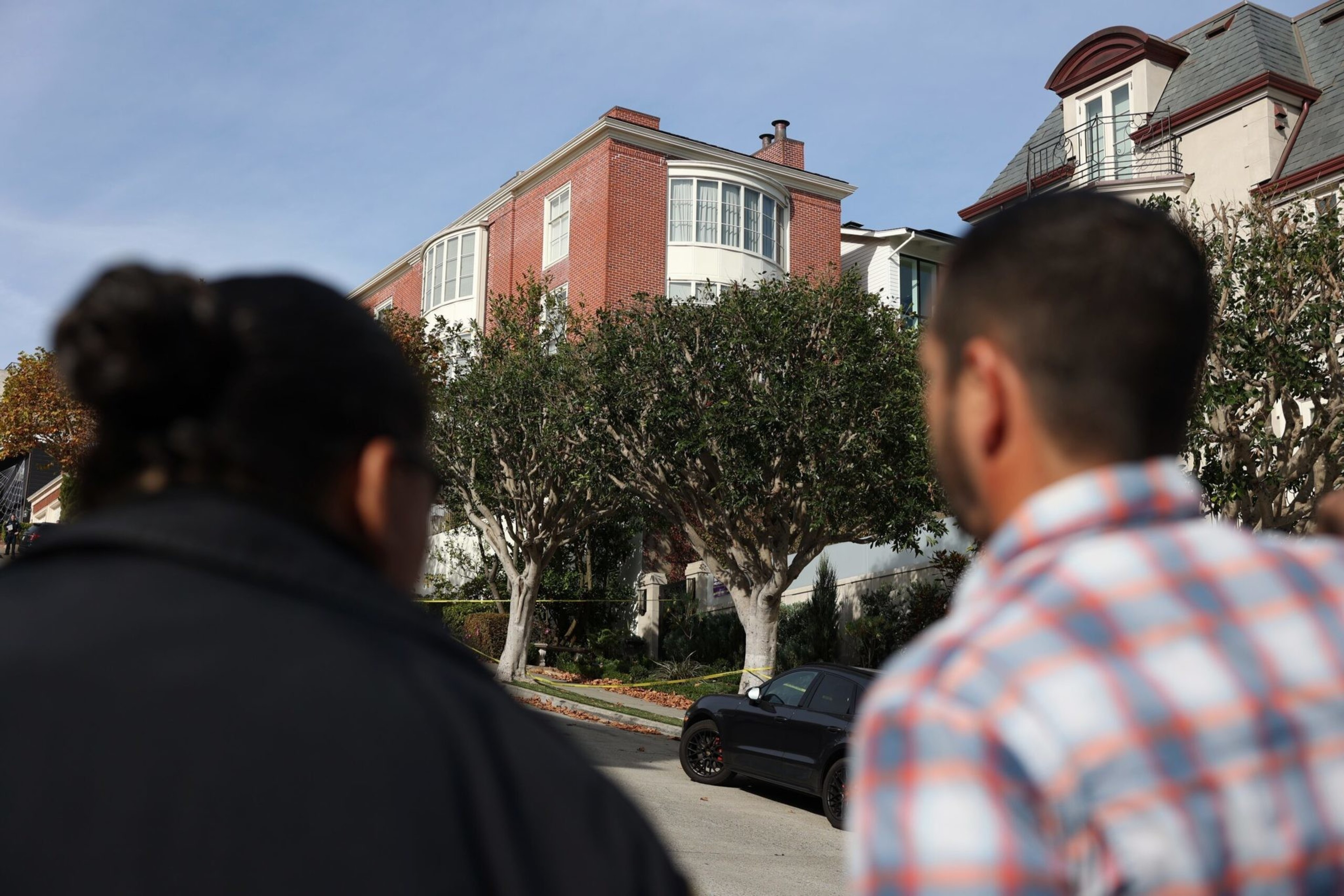 Two people are seen from behind looking at a residential street with large trees, a black car, and multi-story brick and stone houses.