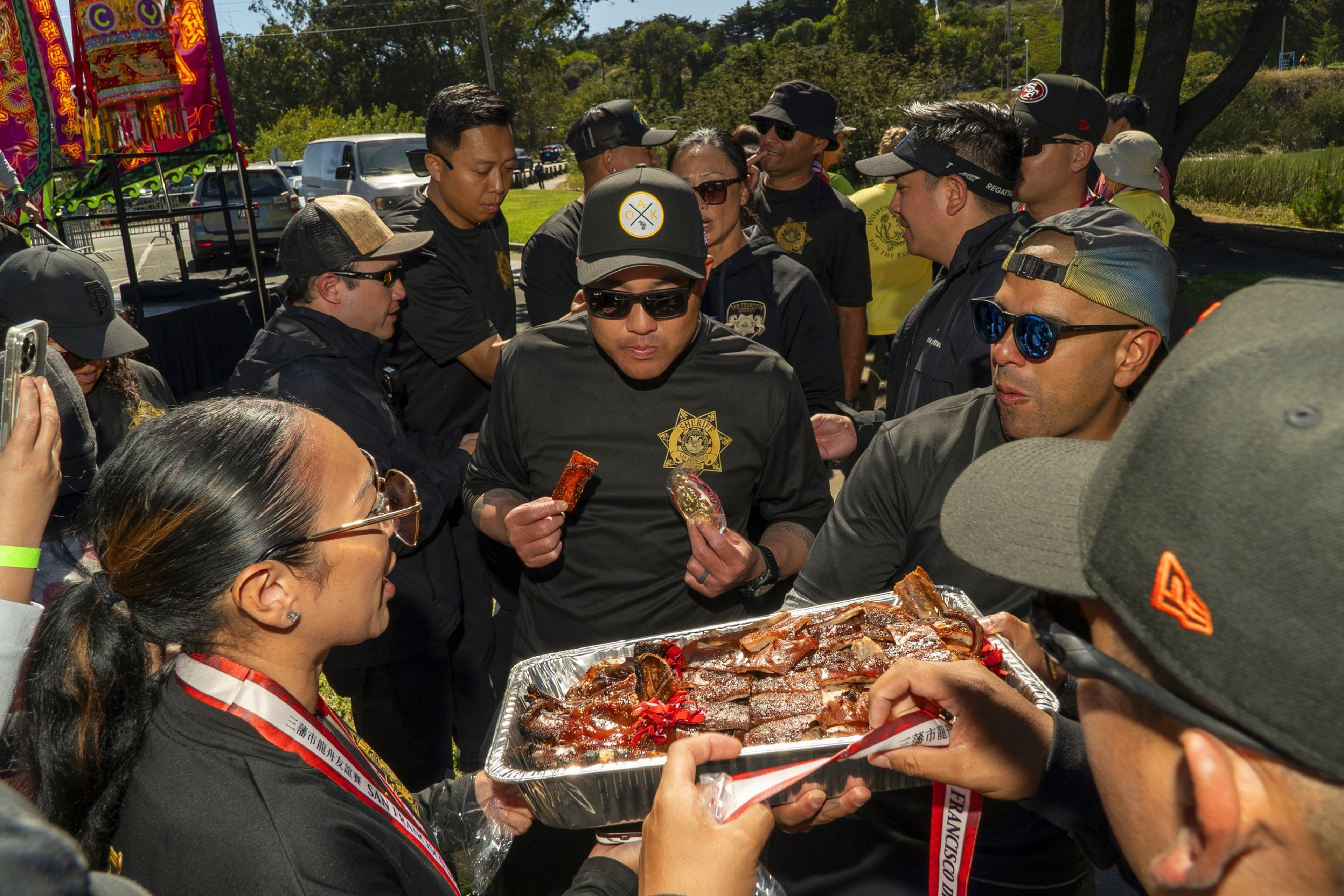 A group of people outdoors, many wearing dark clothing and hats, share a large tray of grilled meat, with one man holding two pieces in his hands.