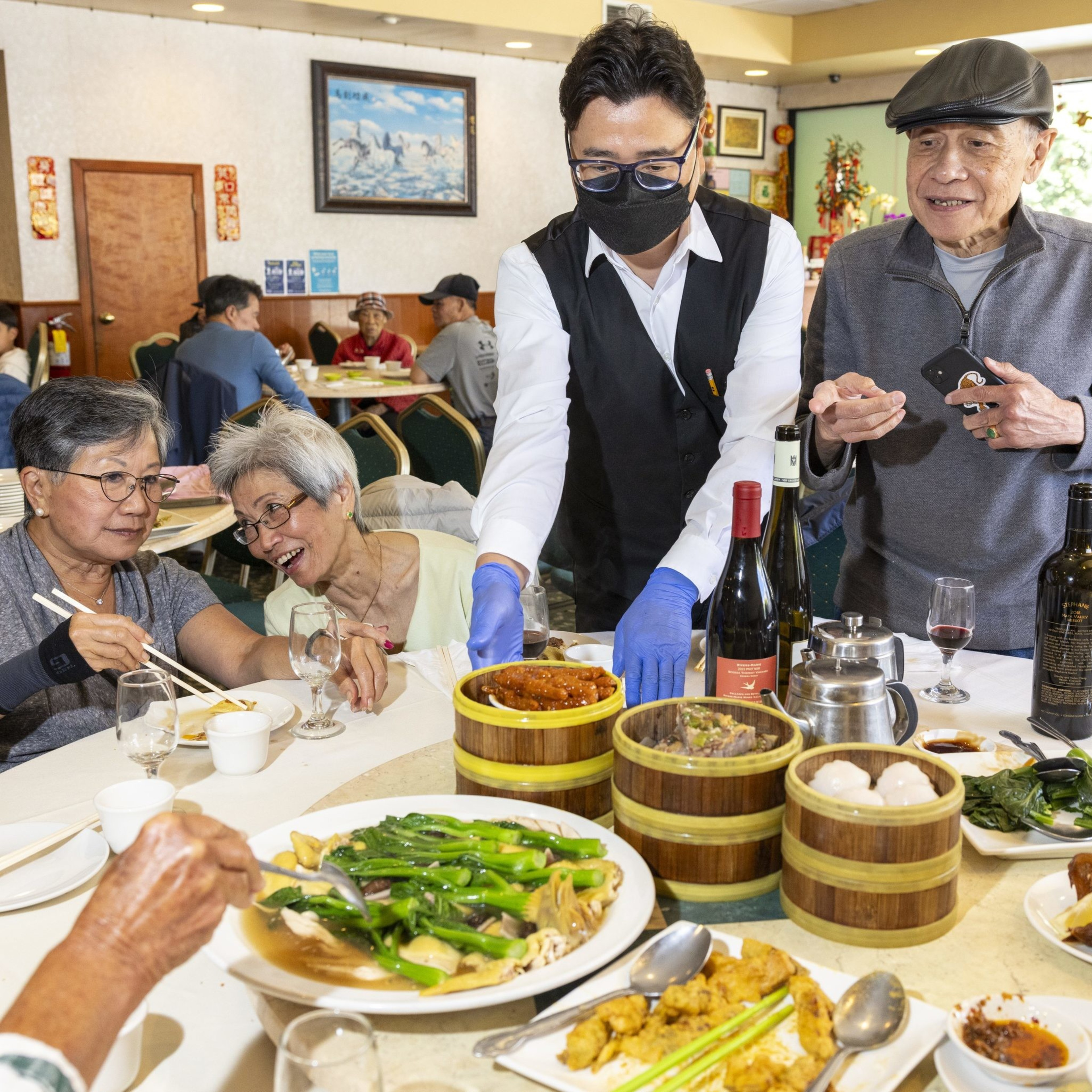 An attentive waiter in a black vest and gloves serves food to a cheerful group of diners in a well-decorated restaurant, with dim sum and wine on the table.