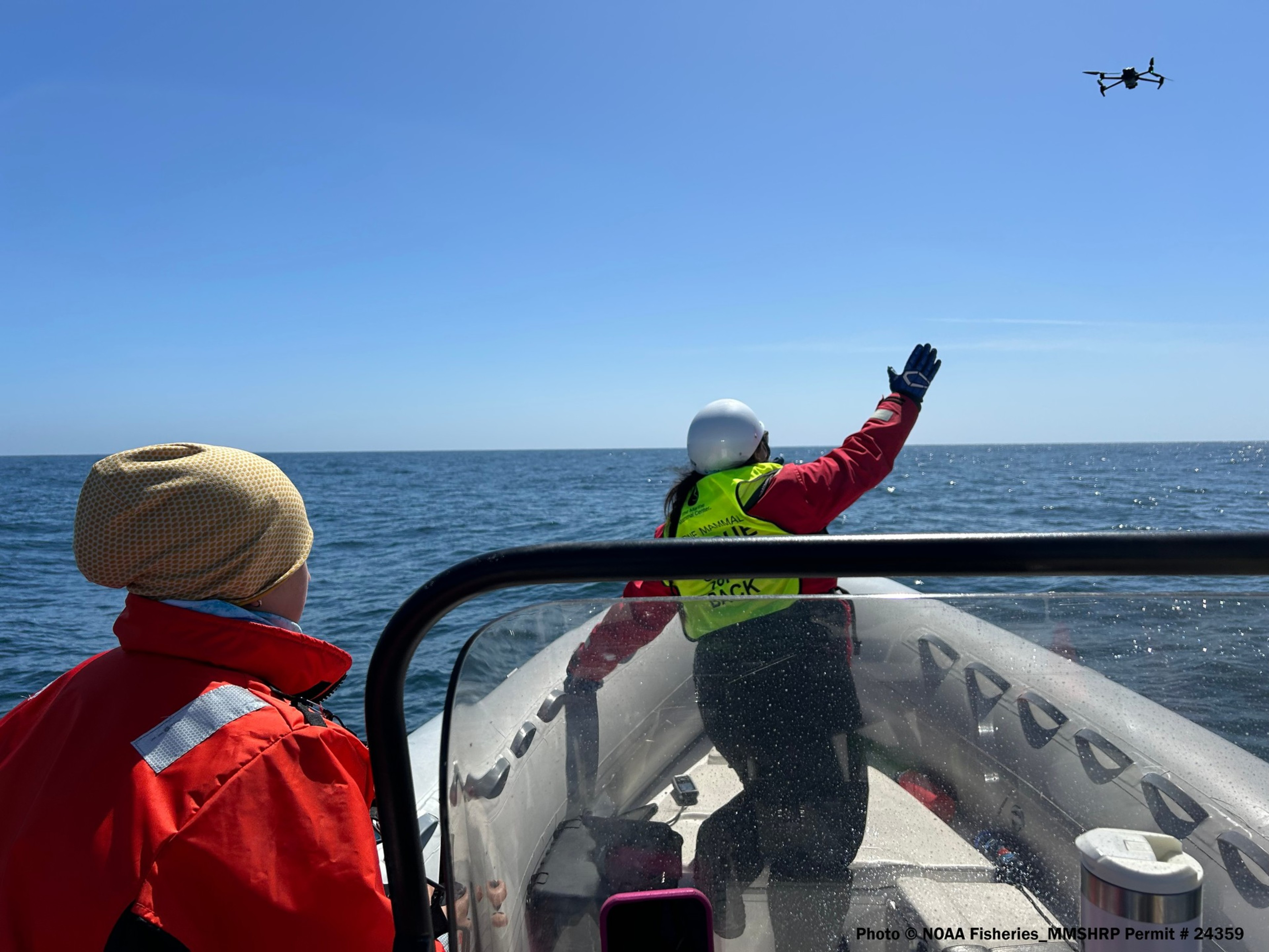 Two people on a boat, one guiding a drone over the sea, with clear skies above.