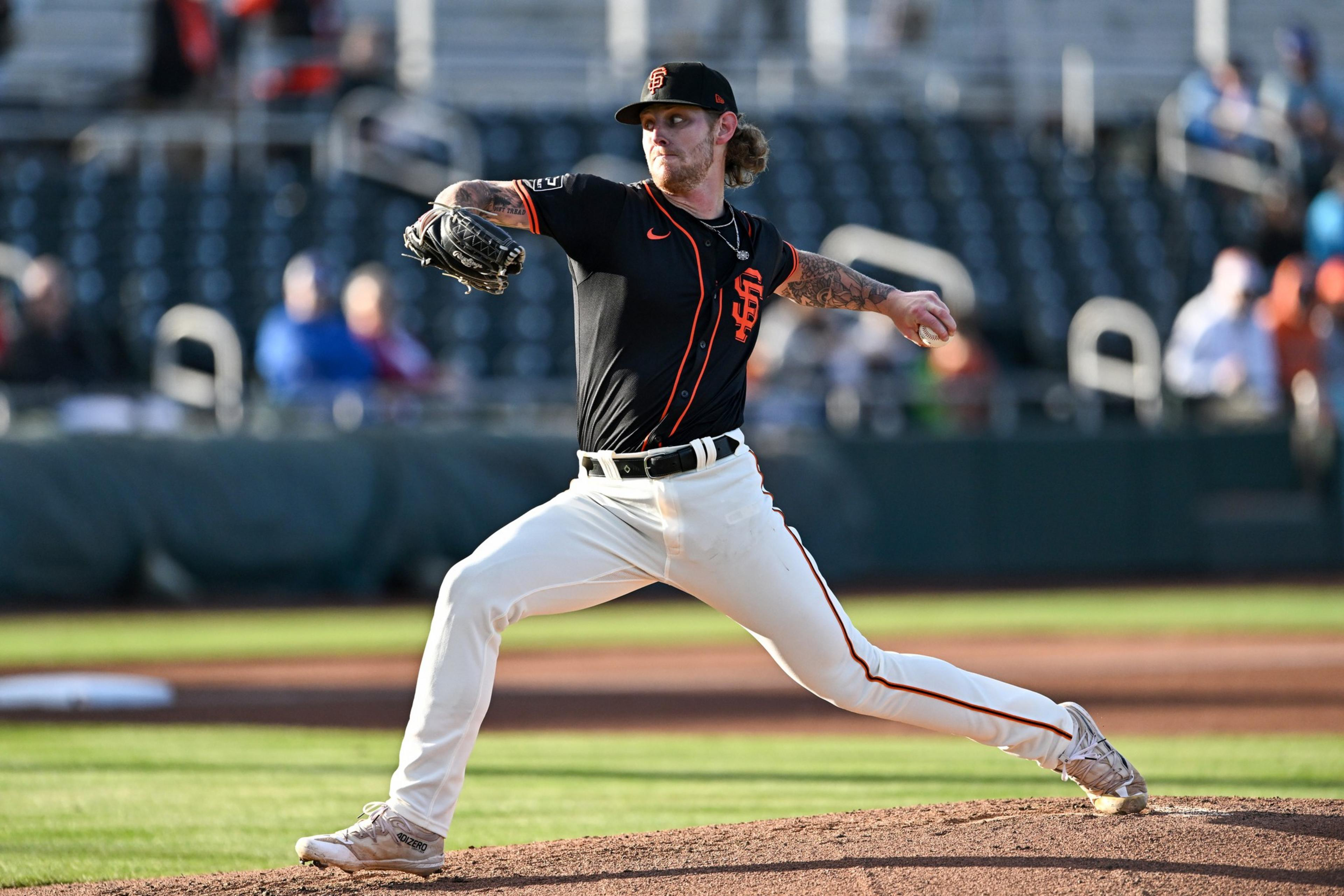 A baseball pitcher in a black and orange uniform launches a pitch on the mound in a stadium. The stands are mostly empty and blurred in the background.