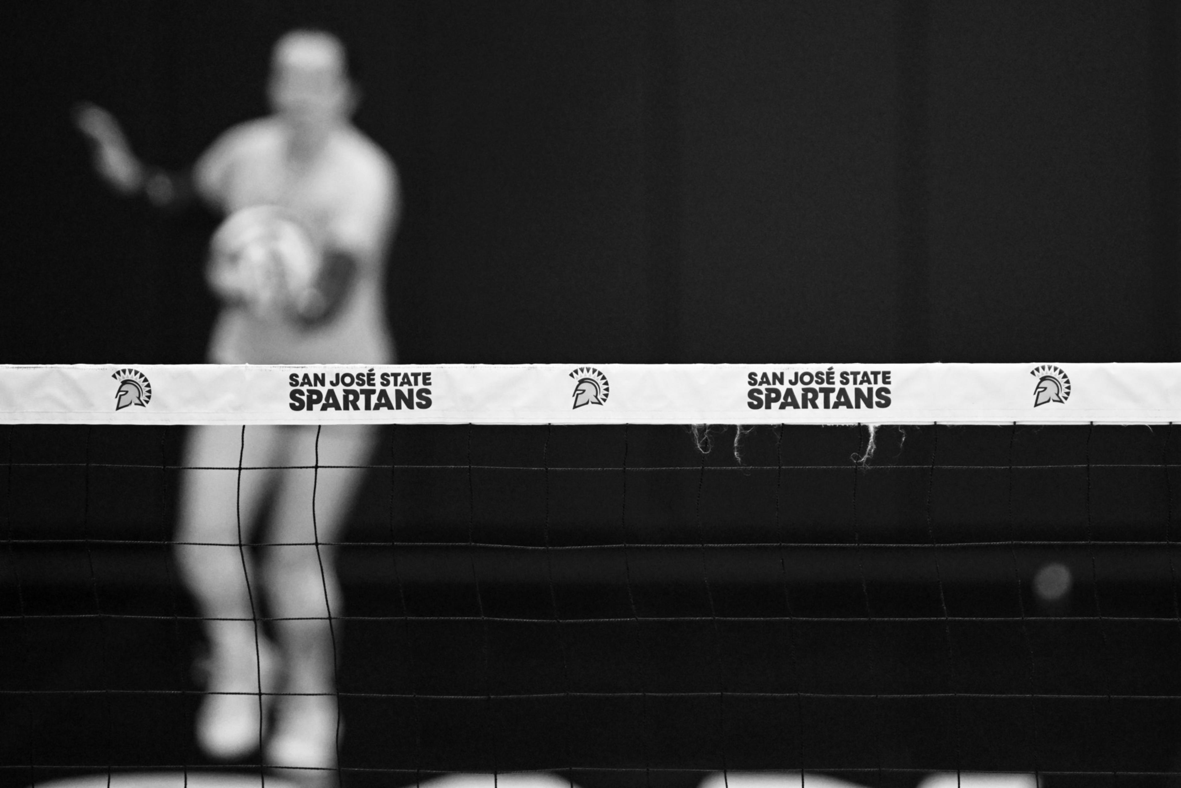 A volleyball net with "San JosÃ© State Spartans" branding is in focus, while a blurred figure in the background appears to be holding a volleyball.