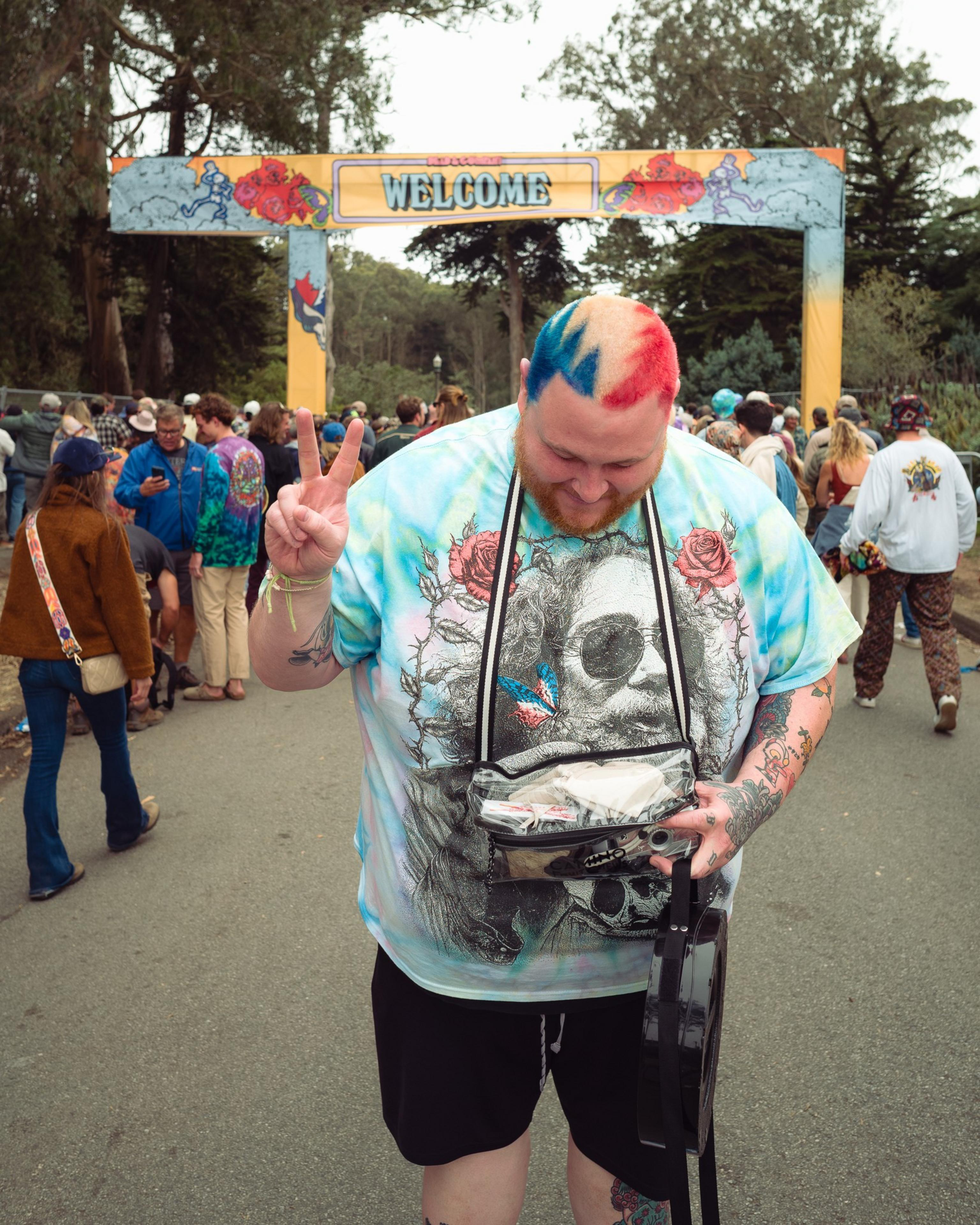 A man with red, blue, and natural hair holds a ukulele and shows a peace sign under a colorful "WELCOME" sign with a crowd behind him.
