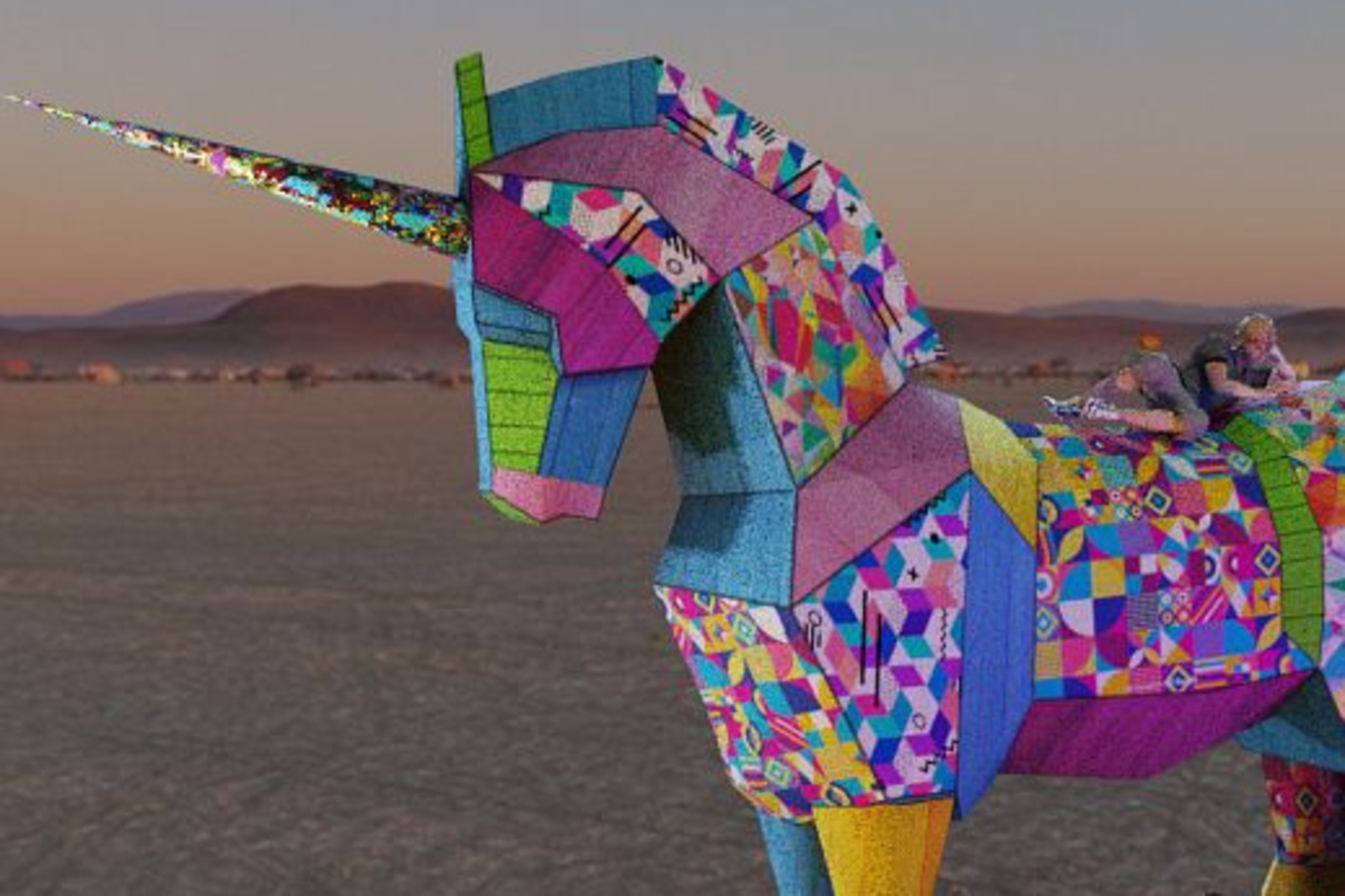 A large, colorful patchwork unicorn sculpture stands on a desert plain with two people lying on its back at sunset.