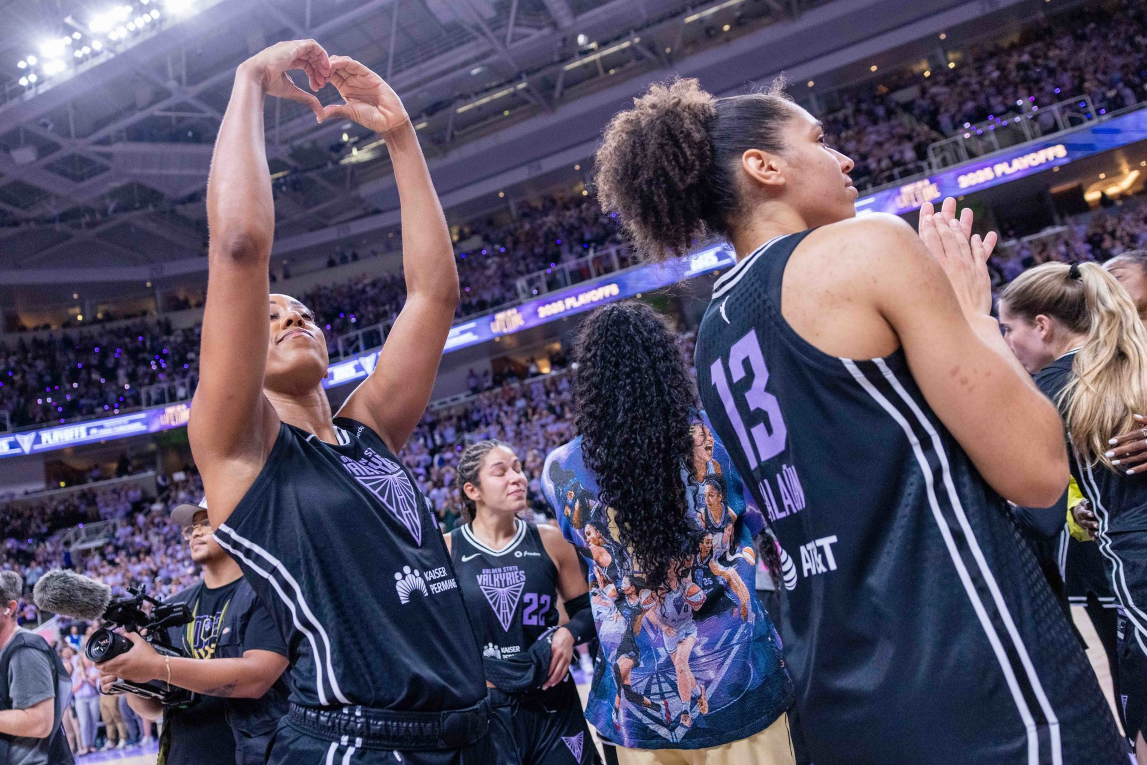 A basketball player forms a heart with her hands while teammates in black jerseys, one numbered 13, celebrate on the court with a crowd in the background.