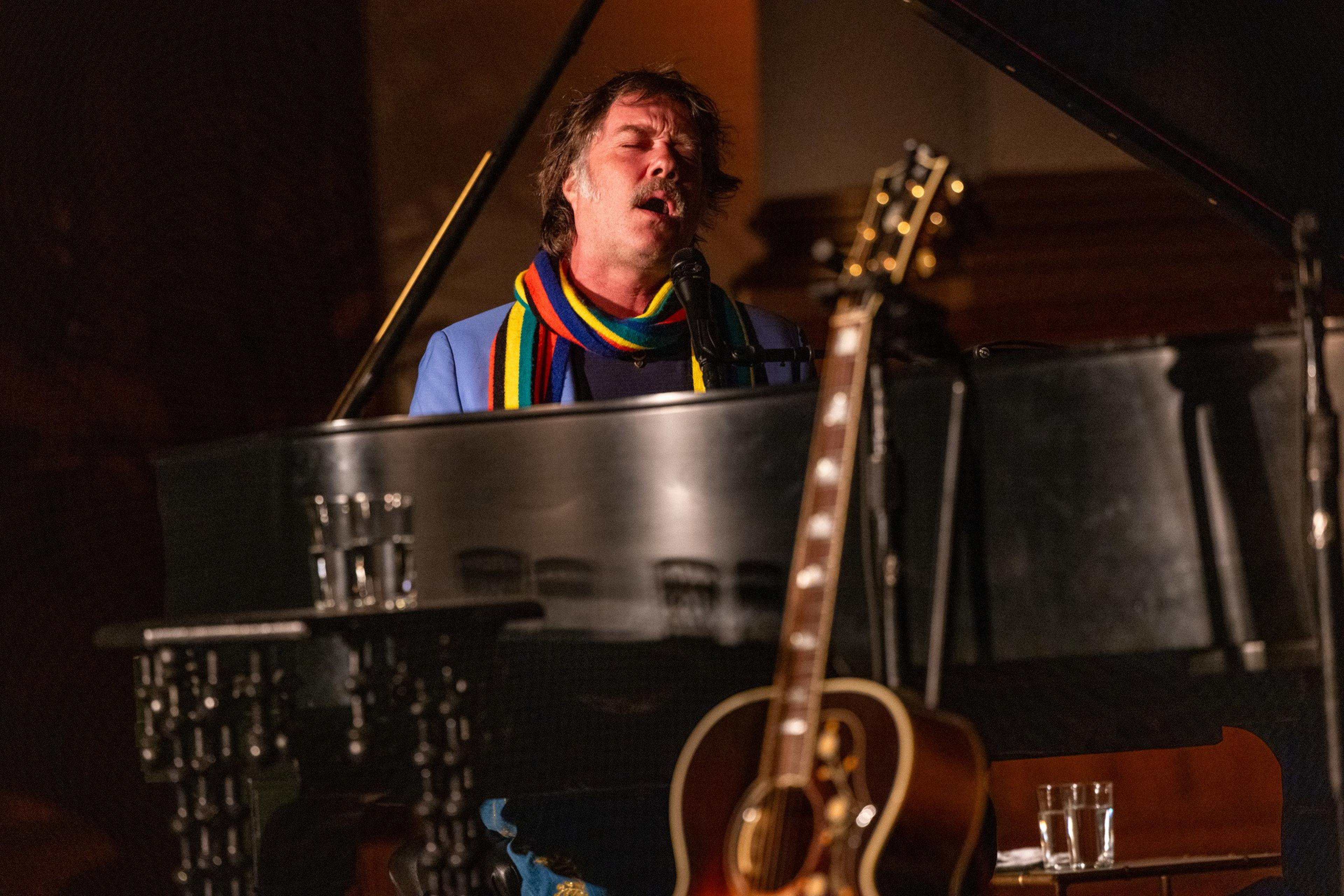 A person passionately sings at a black grand piano, wearing a colorful striped scarf. A guitar leans against the piano, and there are glasses of water nearby.