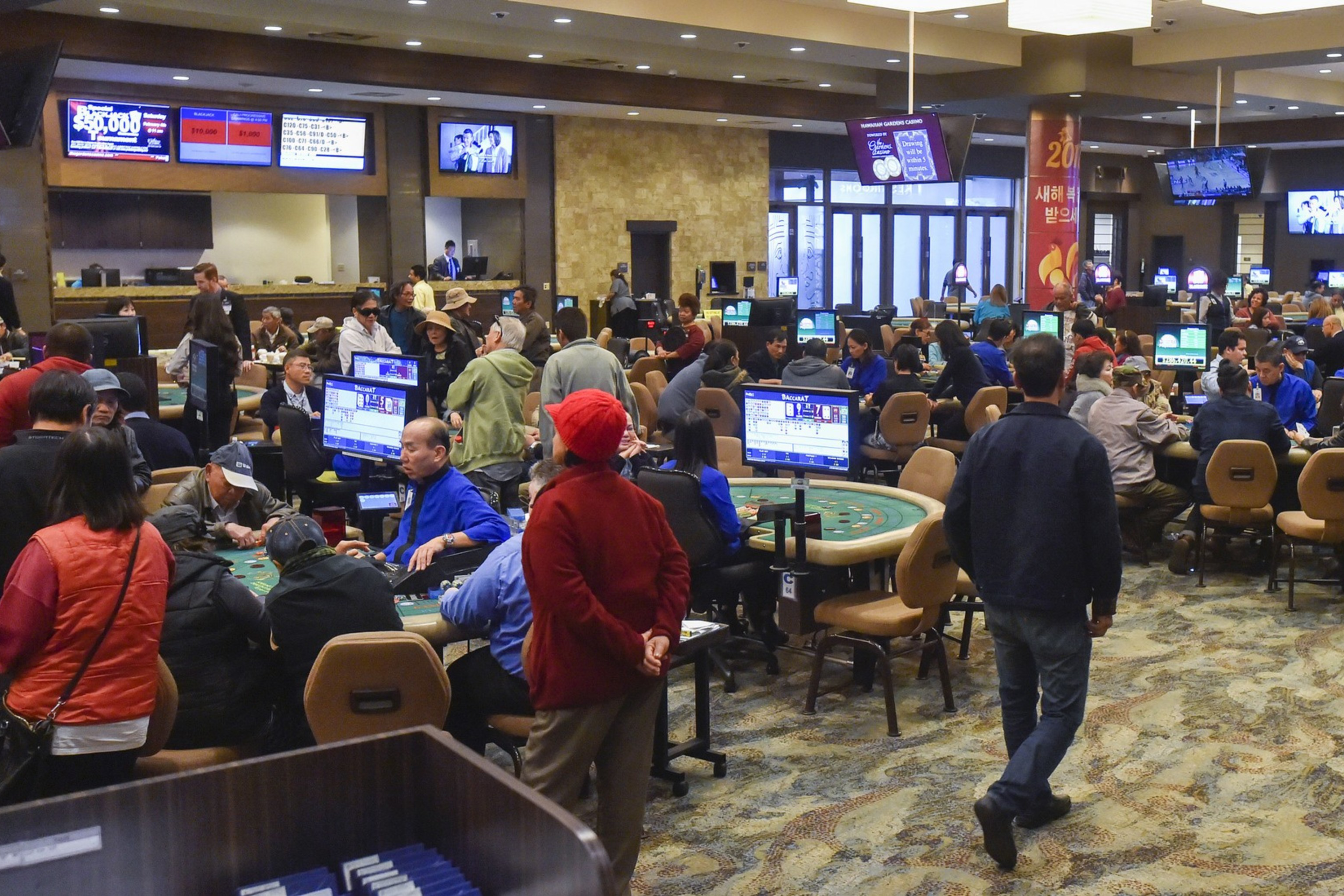 The image shows a busy casino with numerous people sitting and playing games at tables. There are large screens displaying information, and the atmosphere is lively.