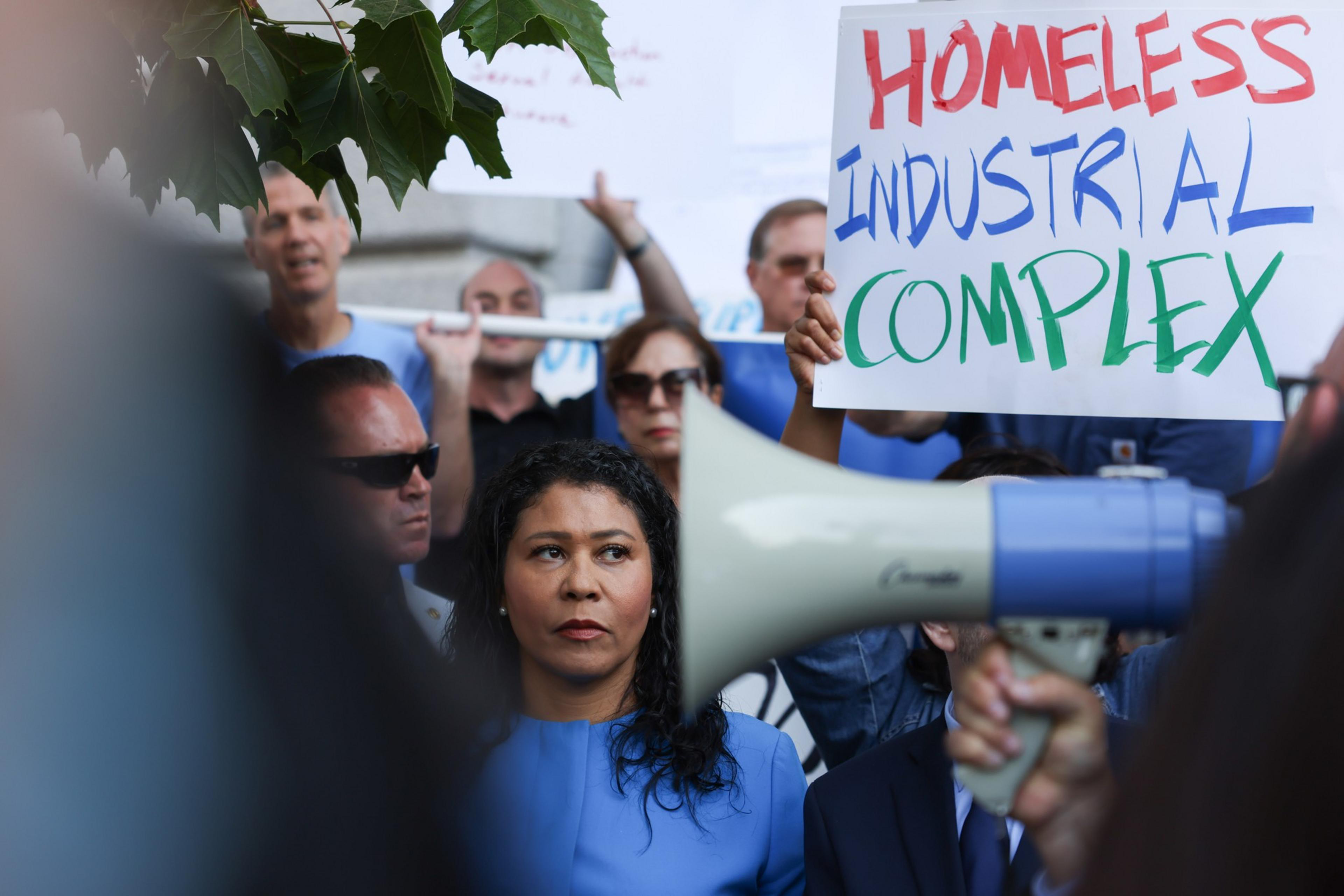 A woman with a megaphone is in focus with protesters and a &quot;HOMELESS INDUSTRIAL COMPLEX&quot; sign in the background.