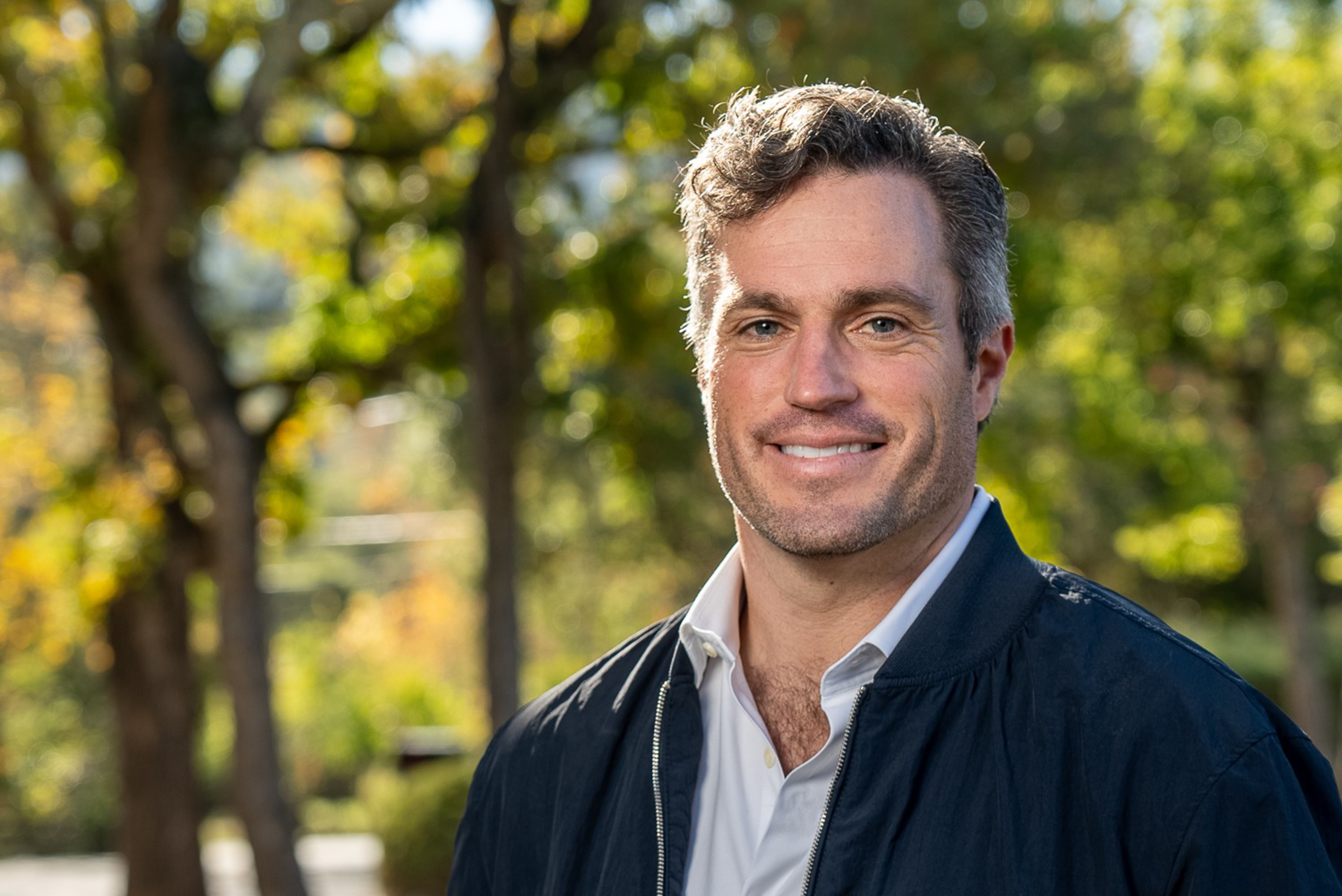 A smiling man with curly hair outdoors, trees with autumn leaves blurred in the background.