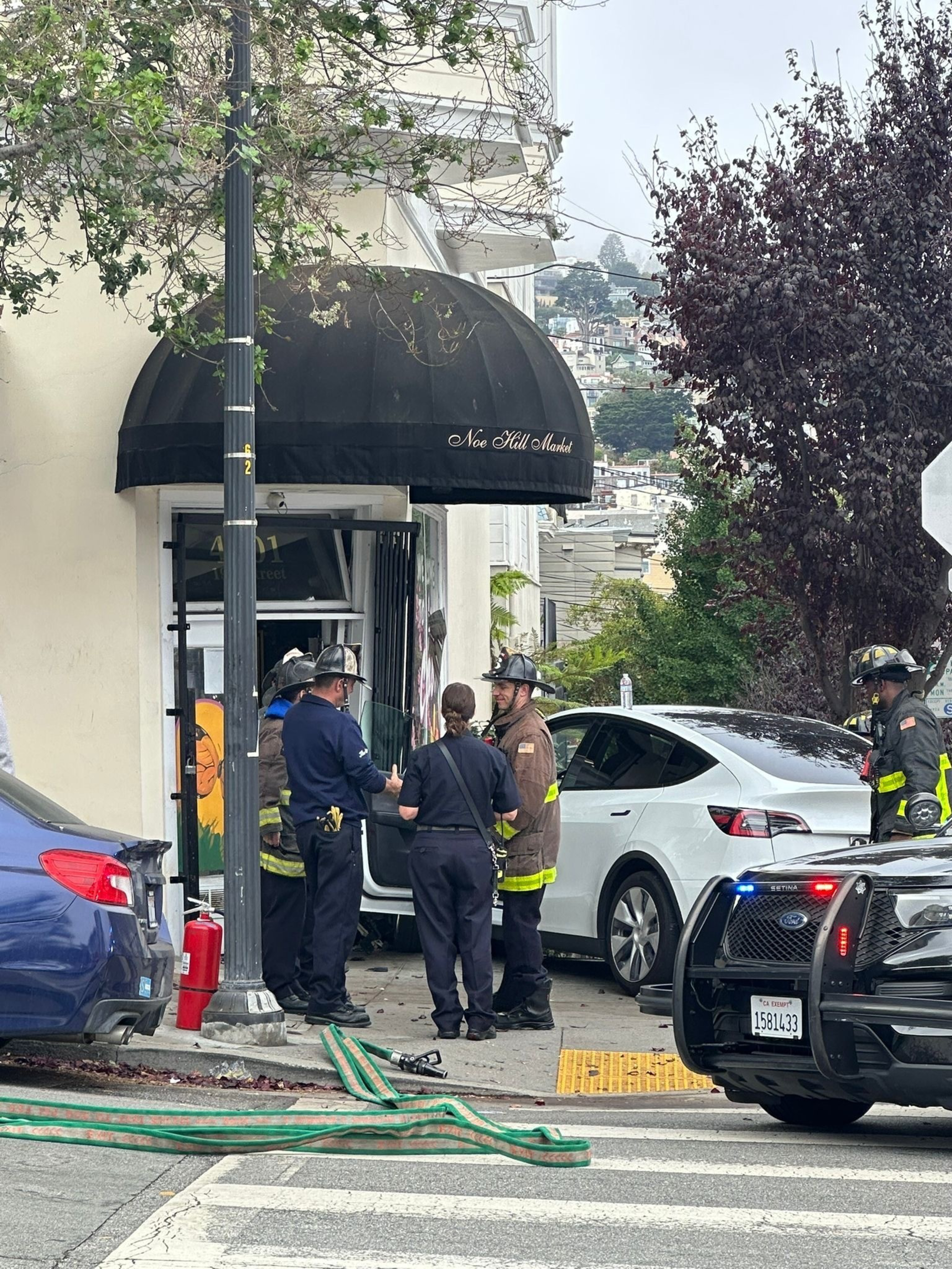 A group of firefighters and a police officer stand outside a store with a black awning, assessing a white car that has crashed into the storefront.