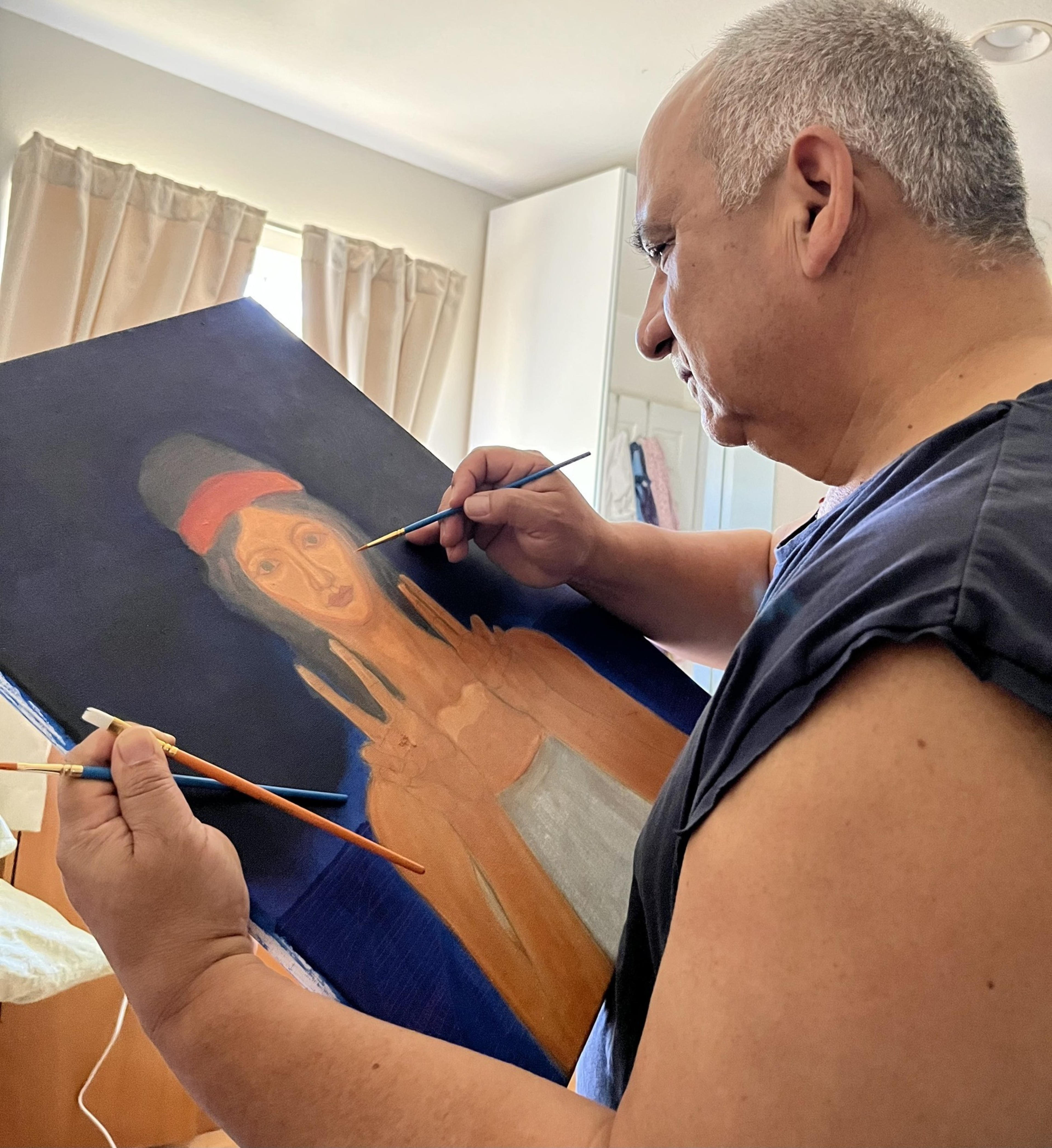 A man with short gray hair concentrates on painting a portrait of a woman wearing a red headband, holding two paintbrushes in a sunlit room.