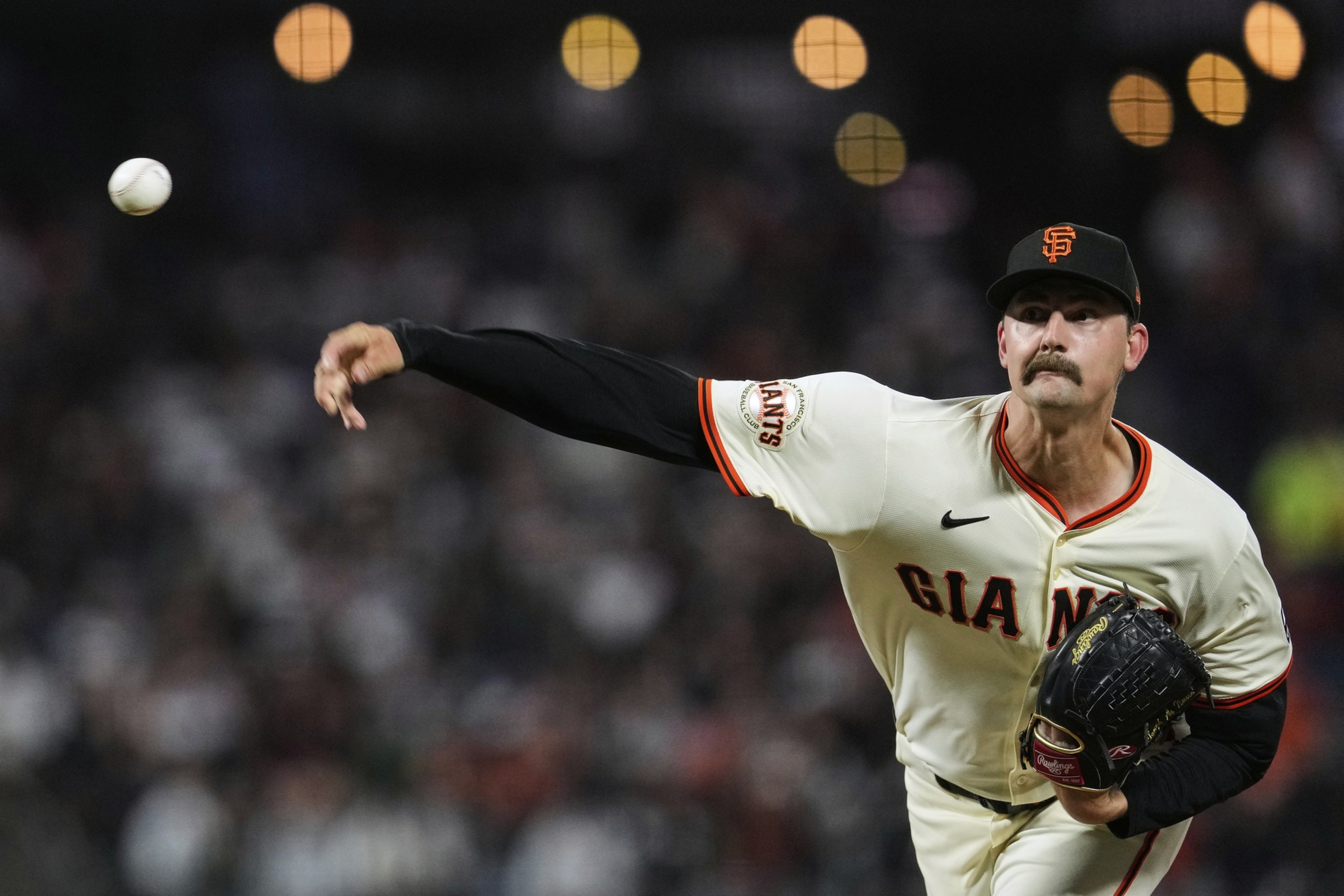 A pitcher in a white Giants uniform with a black glove throws a baseball. The backdrop shows blurred lights and a crowd, creating an action-packed scene.