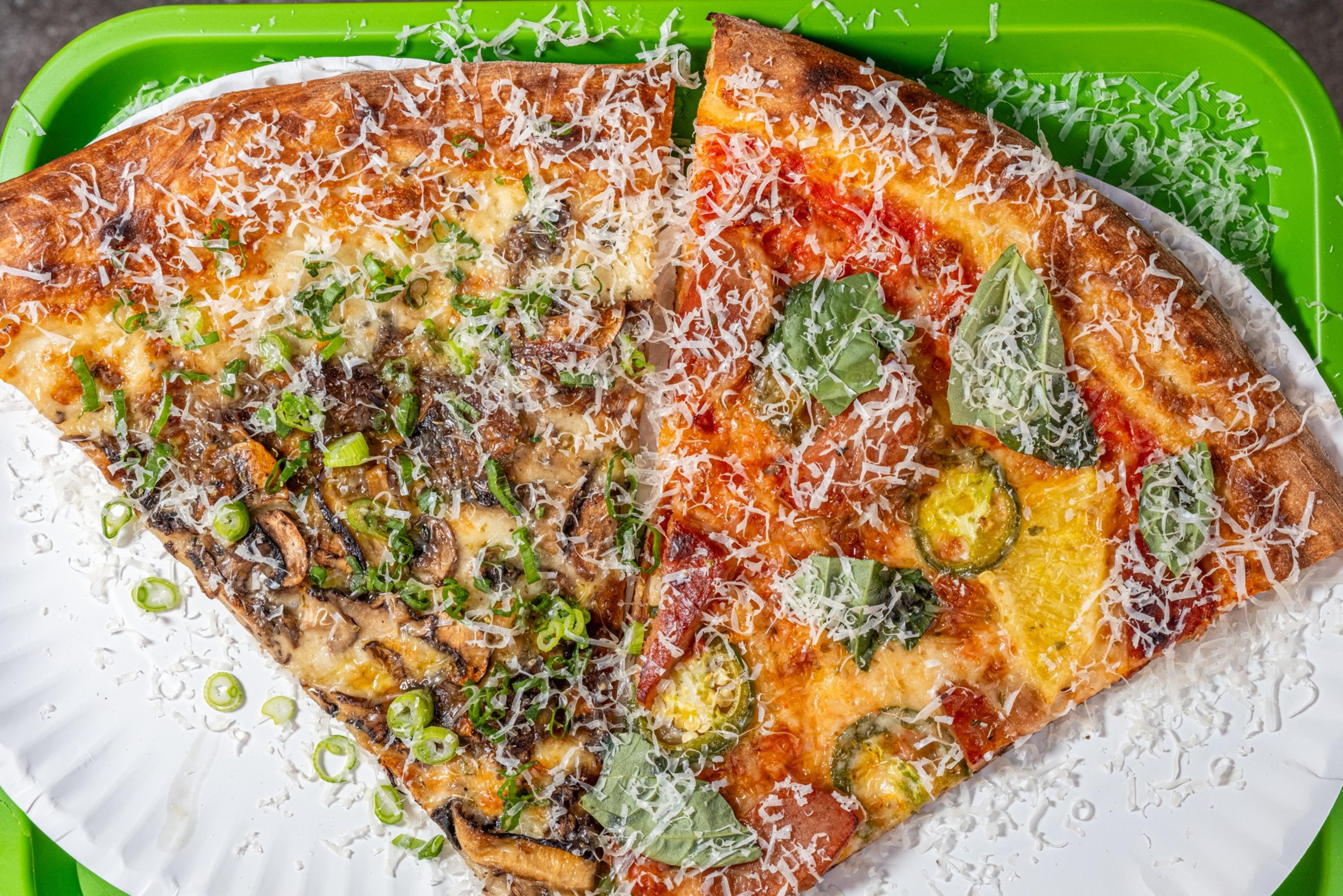 Two pizza slices on a white paper plate, one topped with mushrooms and green onions, the other with basil and jalapeños, both sprinkled with cheese.