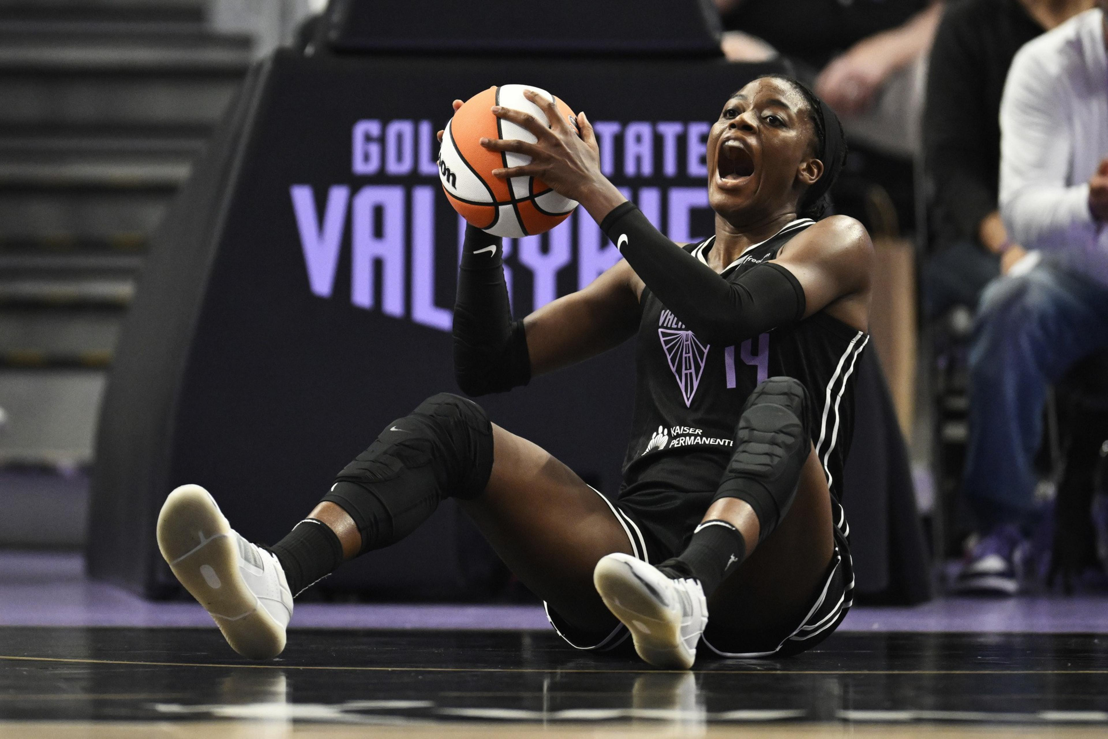 A basketball player wearing a black uniform sits on the court holding a basketball tightly, mouth open as if shouting or reacting.