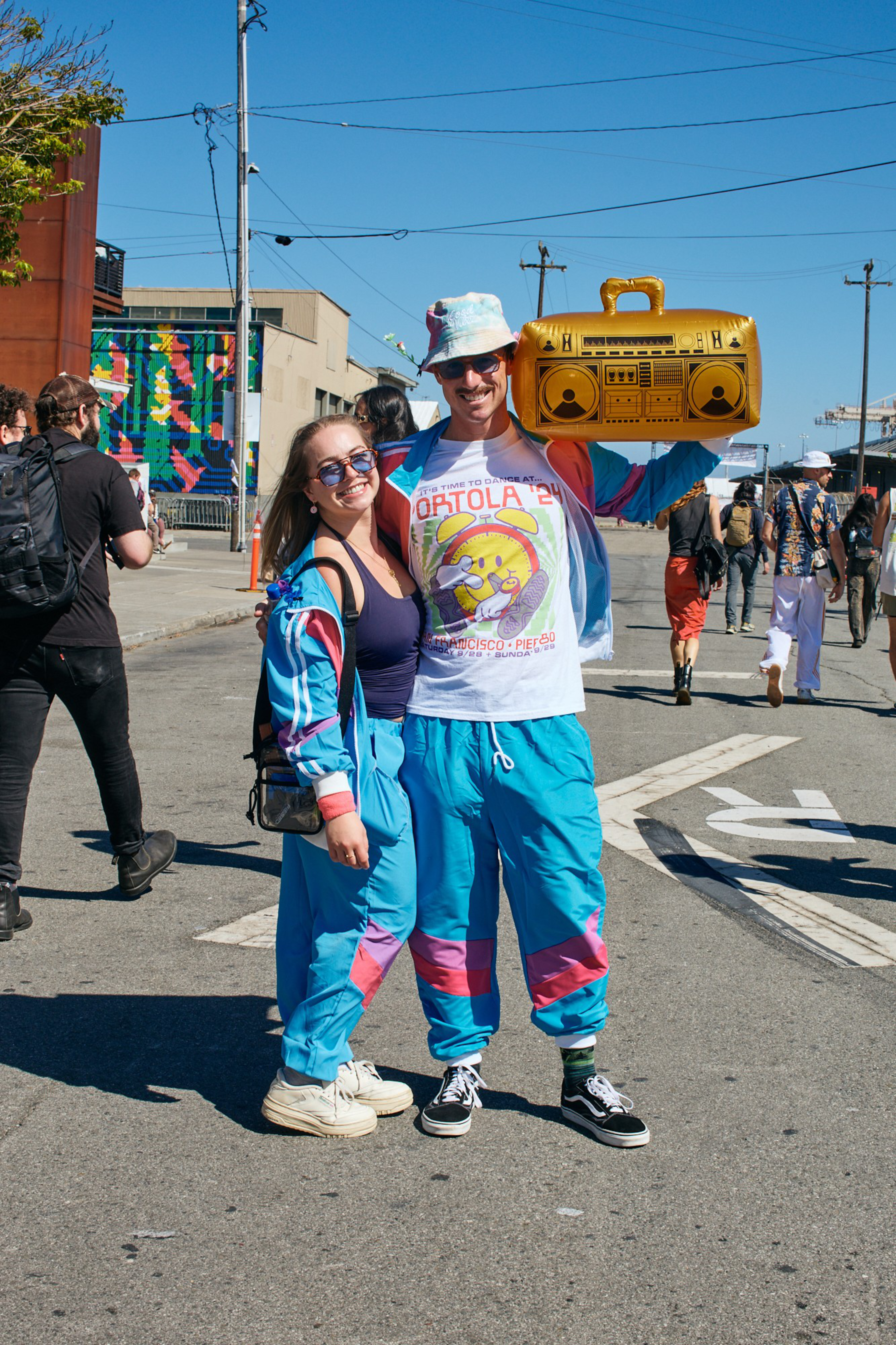 A smiling couple wears matching blue and pink retro tracksuits; the man holds a large gold inflatable boombox over his shoulder.