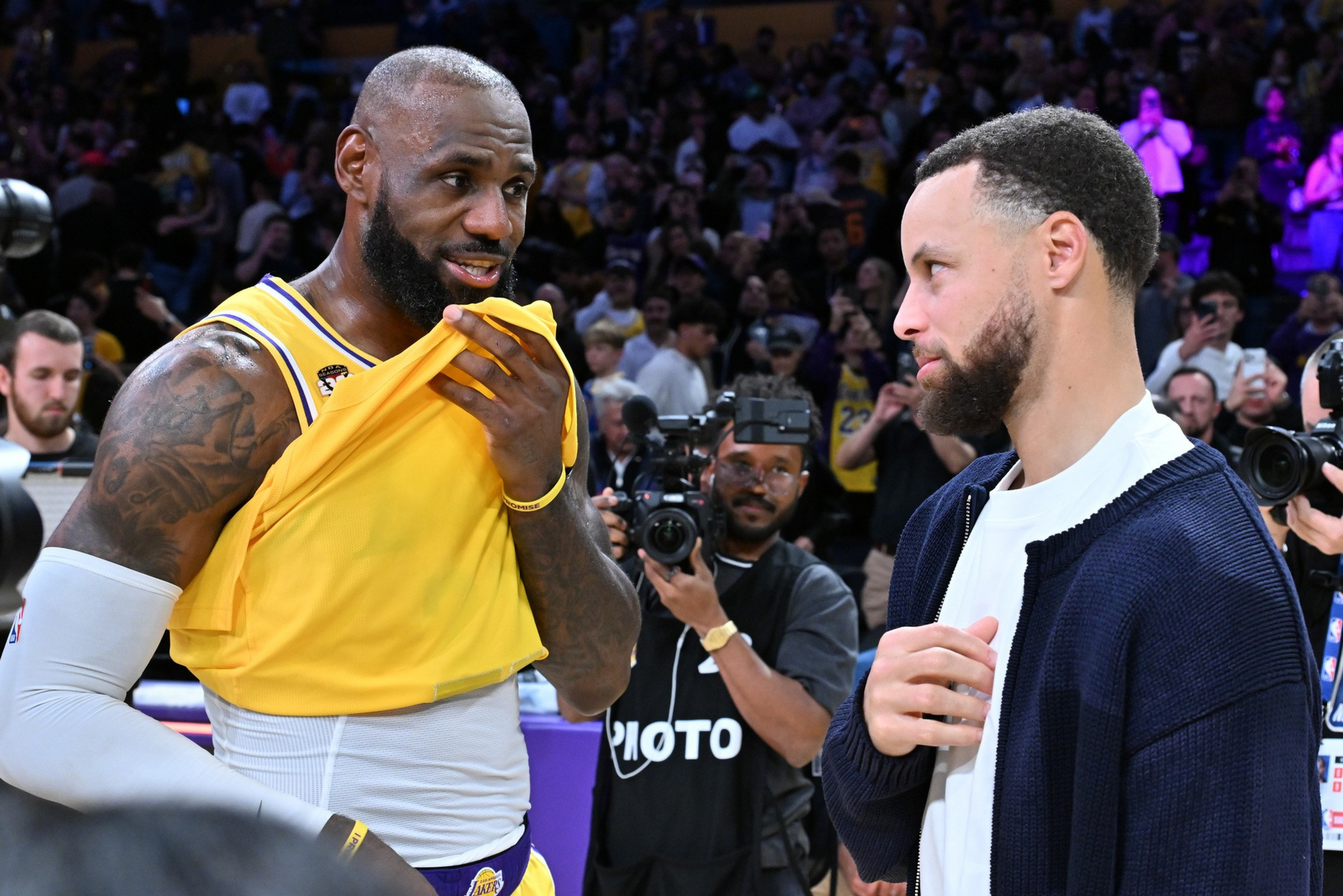 LeBron James in a yellow Lakers jersey talks with Stephen Curry, who wears a white shirt and dark cardigan, while cameras and a crowd capture the moment.