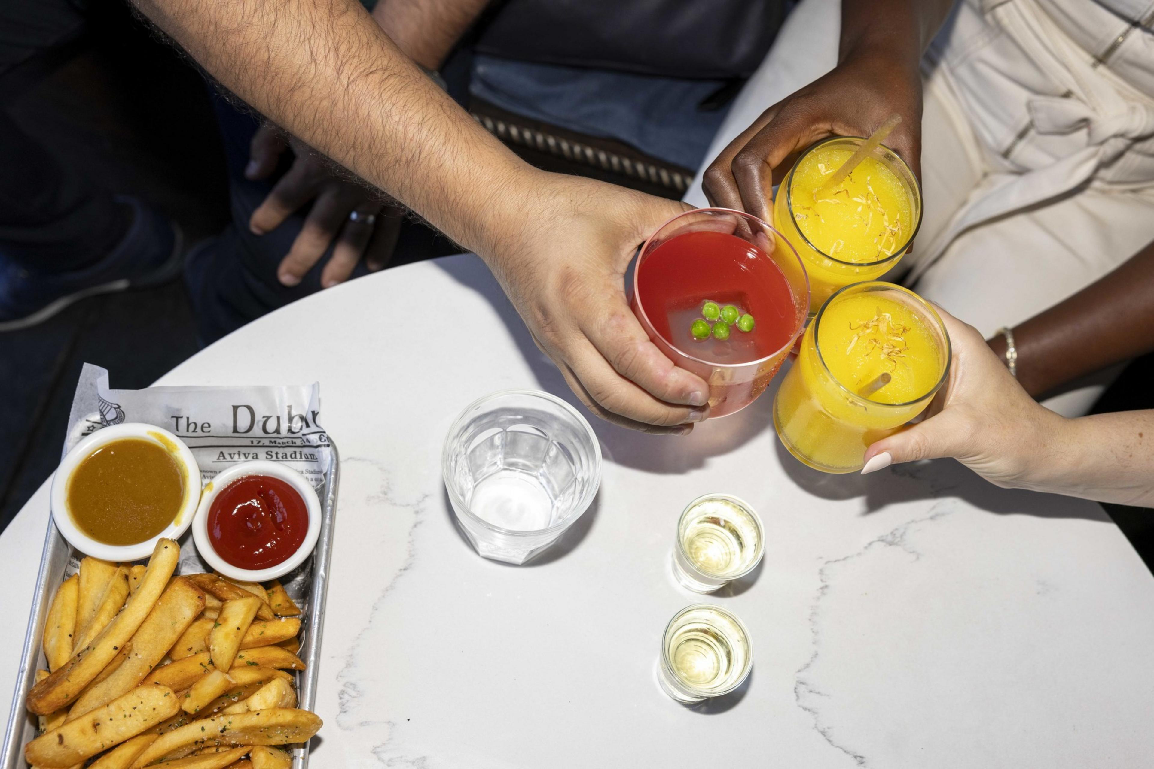 Three hands hold colorful drinks, toasting above a table with fries, ketchup, mustard, a glass of water, and two small shot glasses.