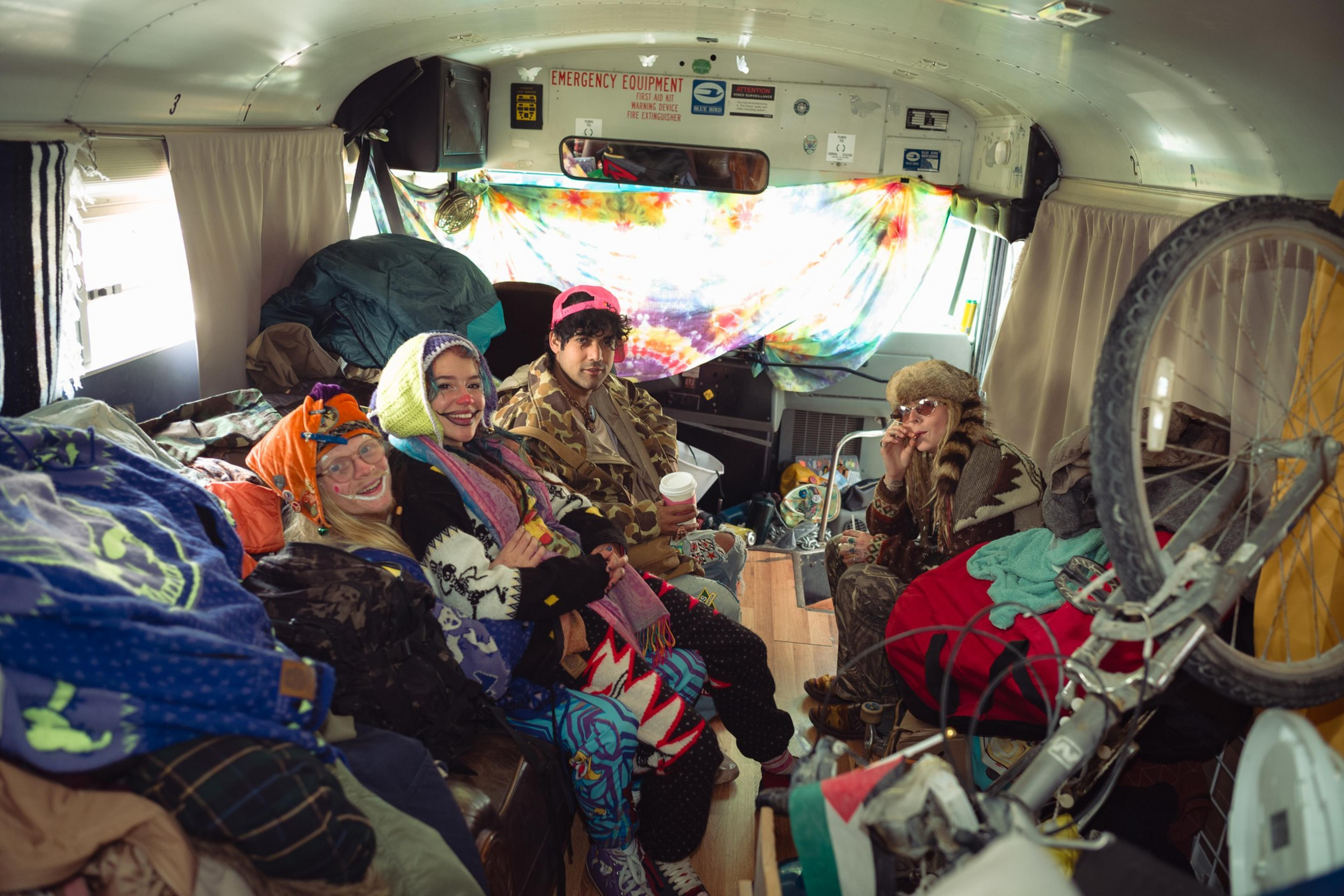 Four people dressed in colorful, eclectic clothing sit inside a cluttered bus with a tie-dye curtain covering the windshield.