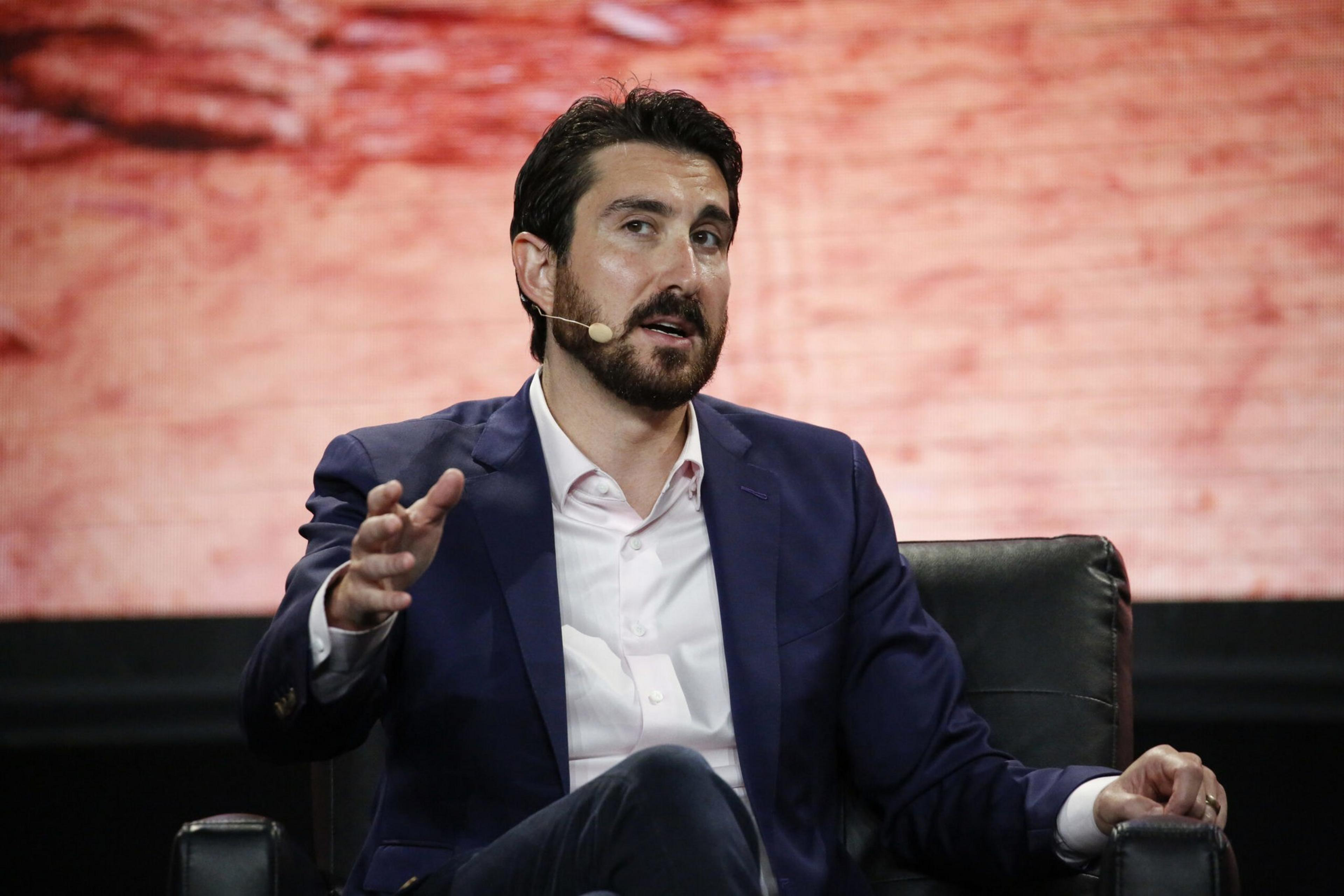 A man with dark hair and beard wearing a navy blue suit and white shirt speaks while seated on a black chair, gesturing with his right hand.