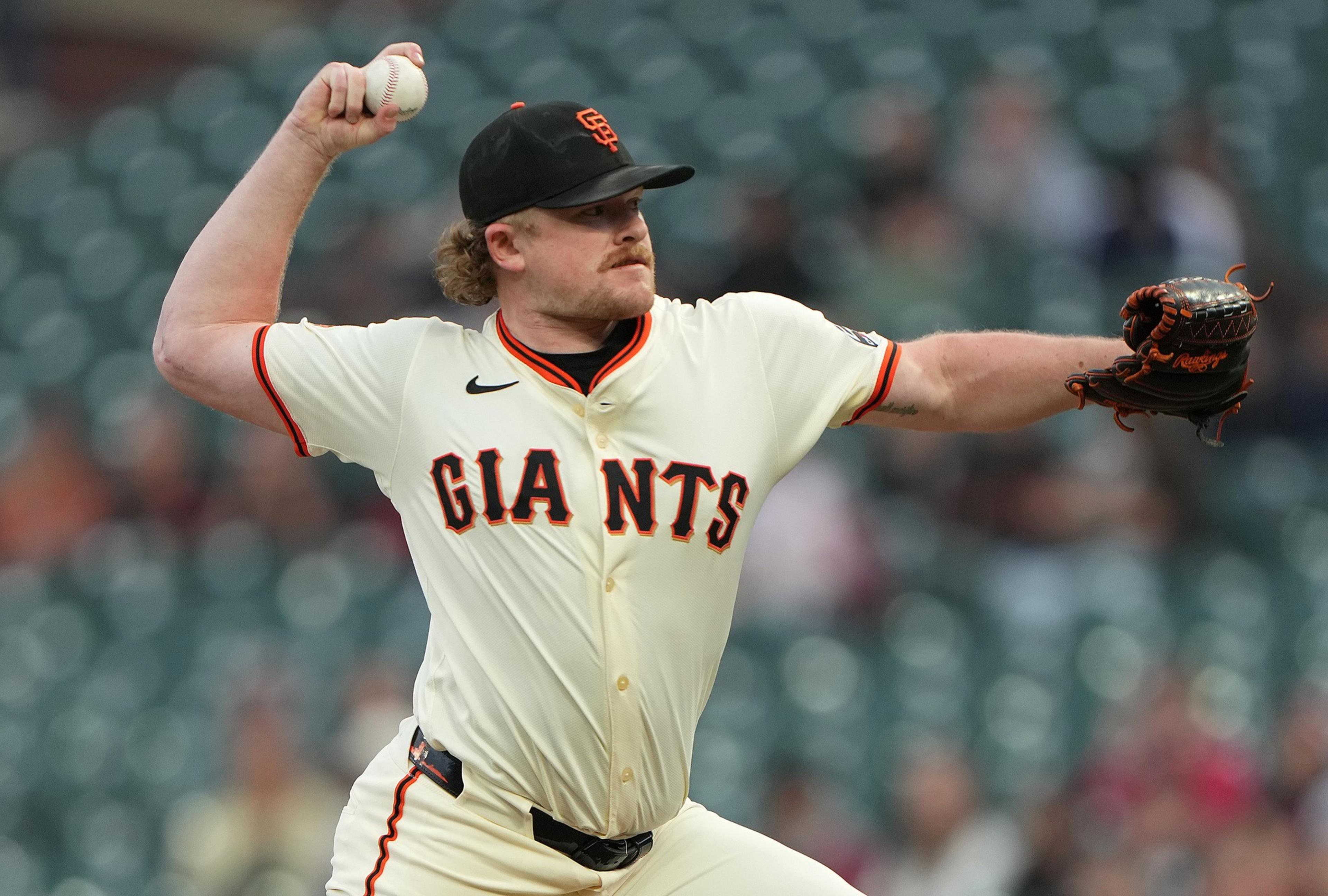 A San Francisco Giants pitcher in a white uniform is mid-throw, gripping a baseball and wearing a black glove on the opposite hand in a blurred stadium.