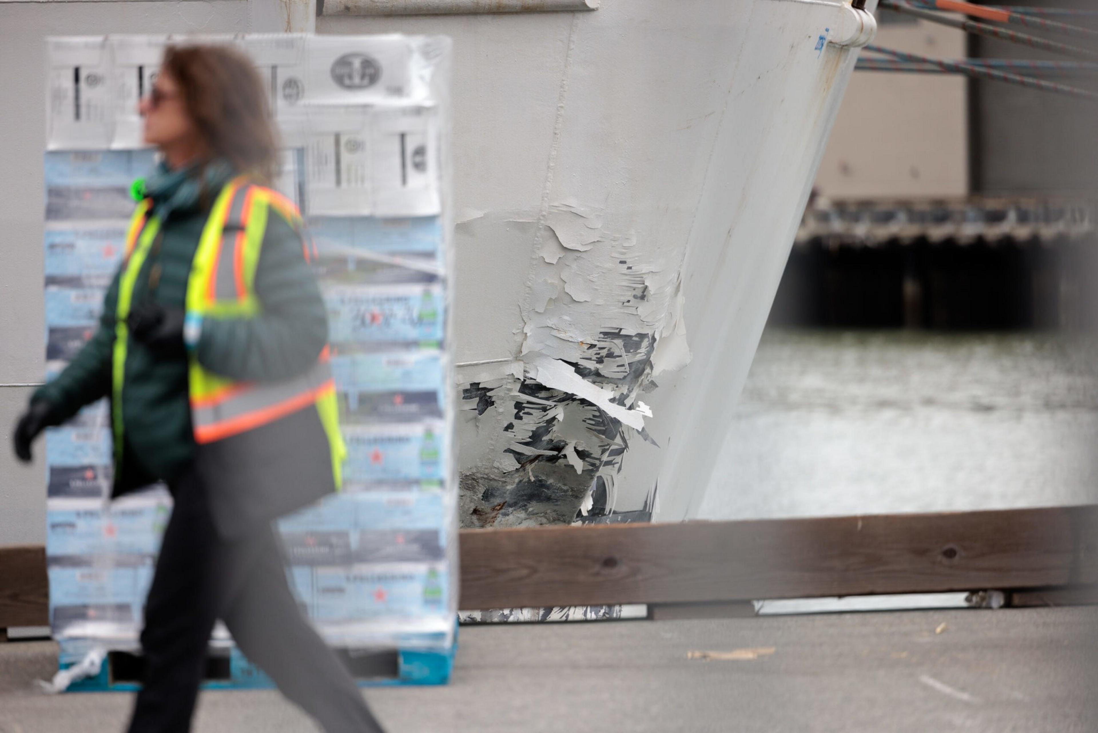 A large ship hull shows visible scraped and damaged paint near the waterline, while a person in a safety vest walks by carrying a bag.
