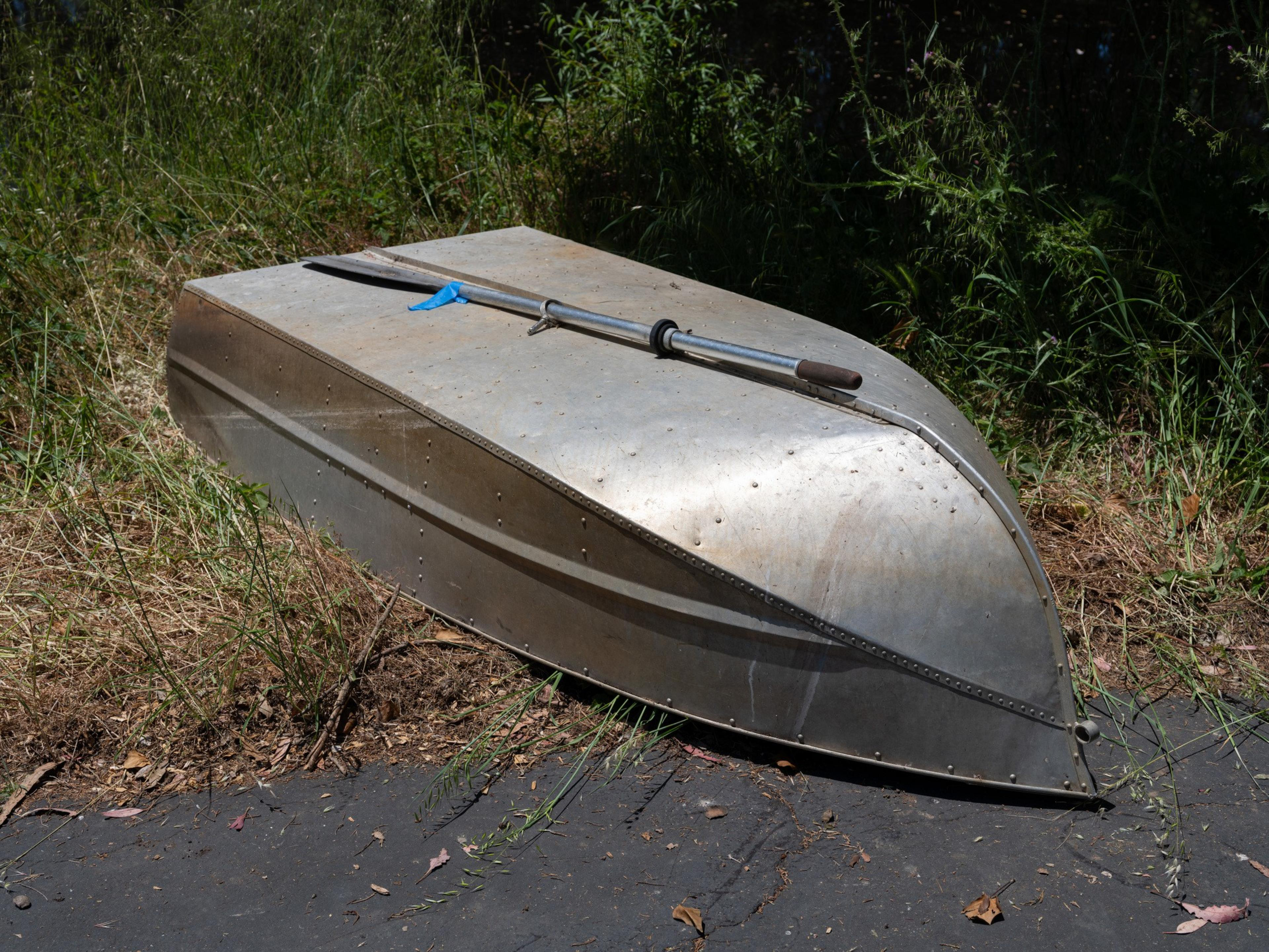 A small, inverted metal rowboat lies on the ground amid grass and leaves, with an oar placed atop it diagonally.