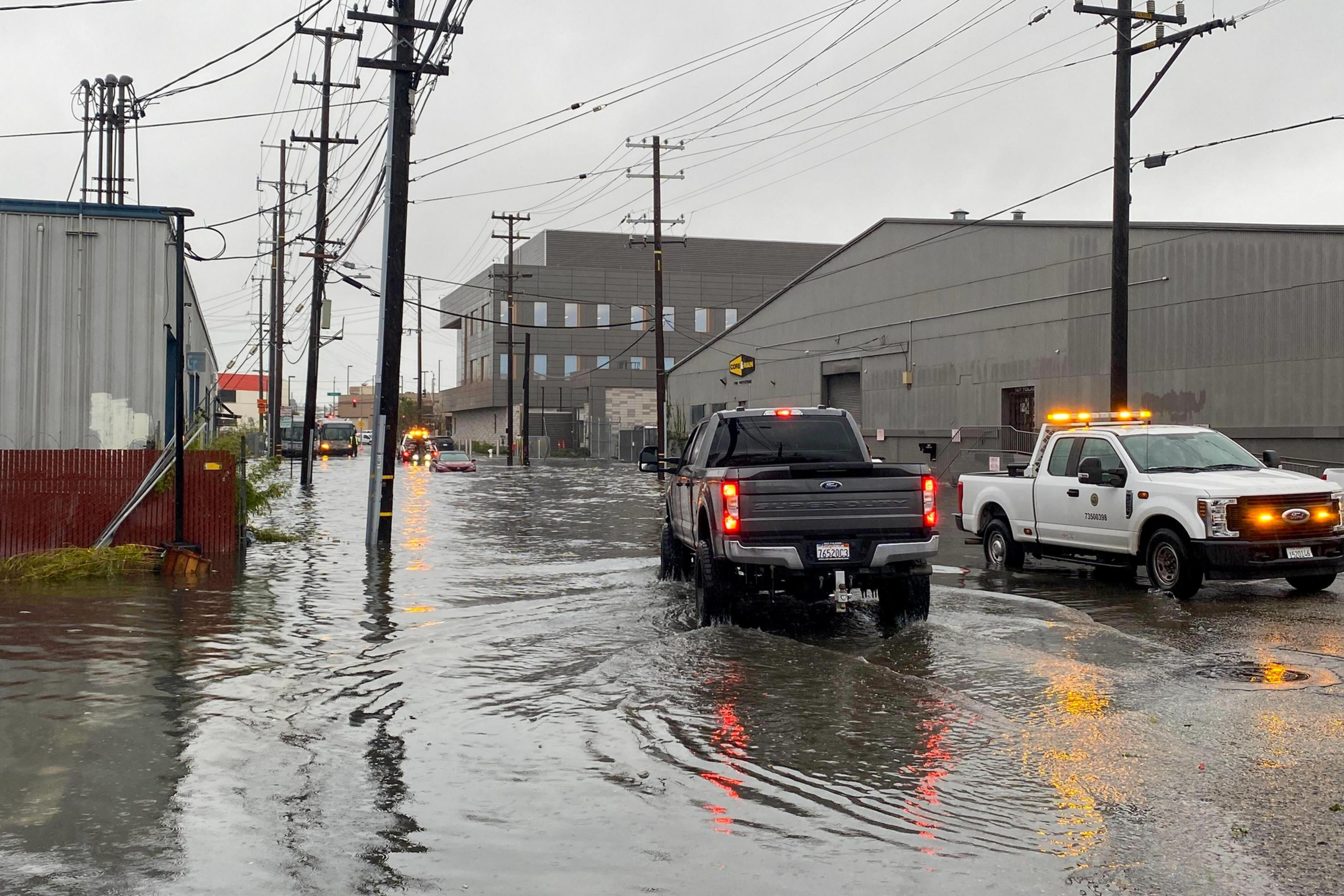 A flooded industrial street with two trucks driving through deep water, utility poles, and buildings under a gray, rainy sky.