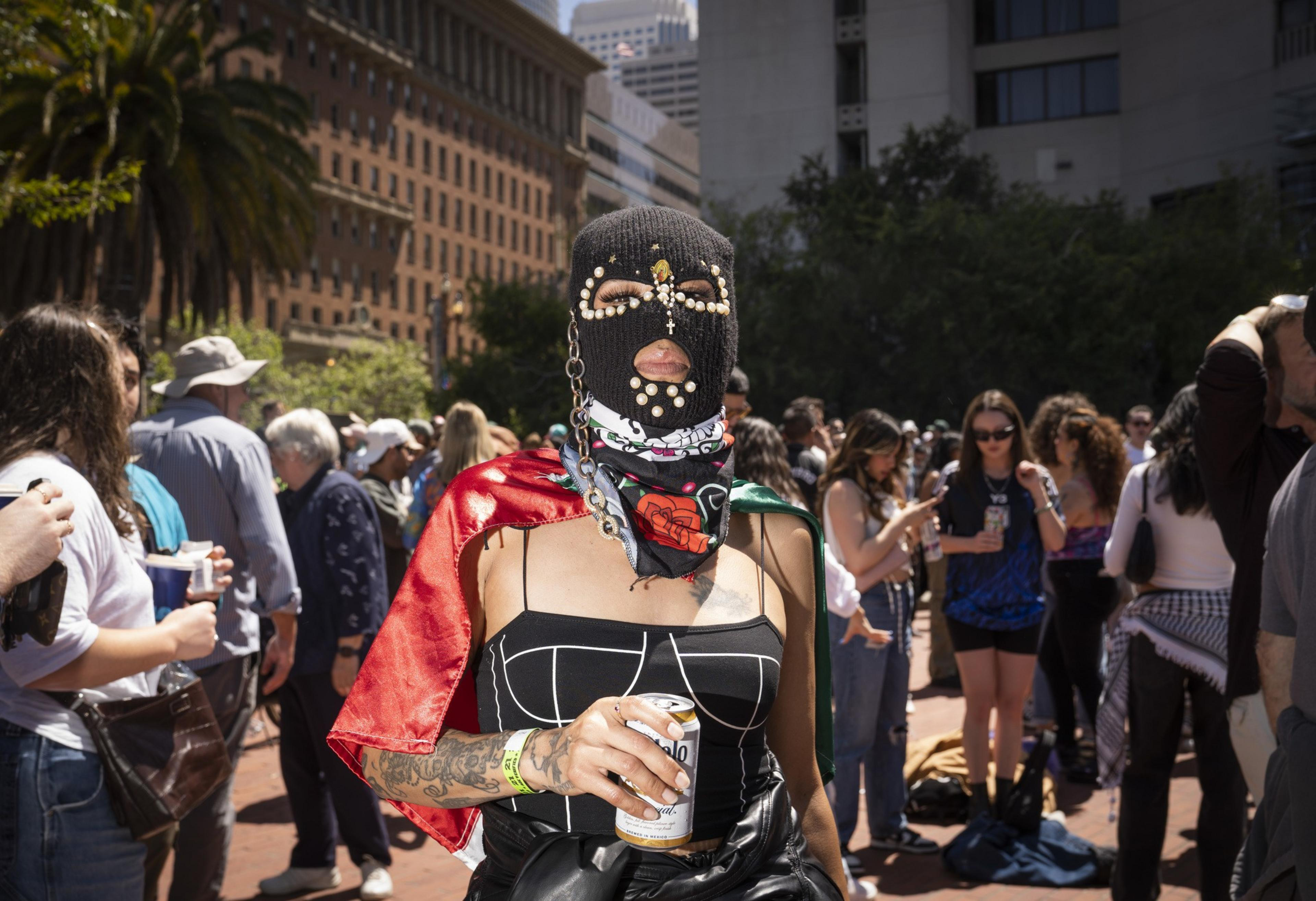 A person wearing a decorated black ski mask, a colorful cape, and a strapless top holds a can in a bustling outdoor crowd with tall buildings in the background.